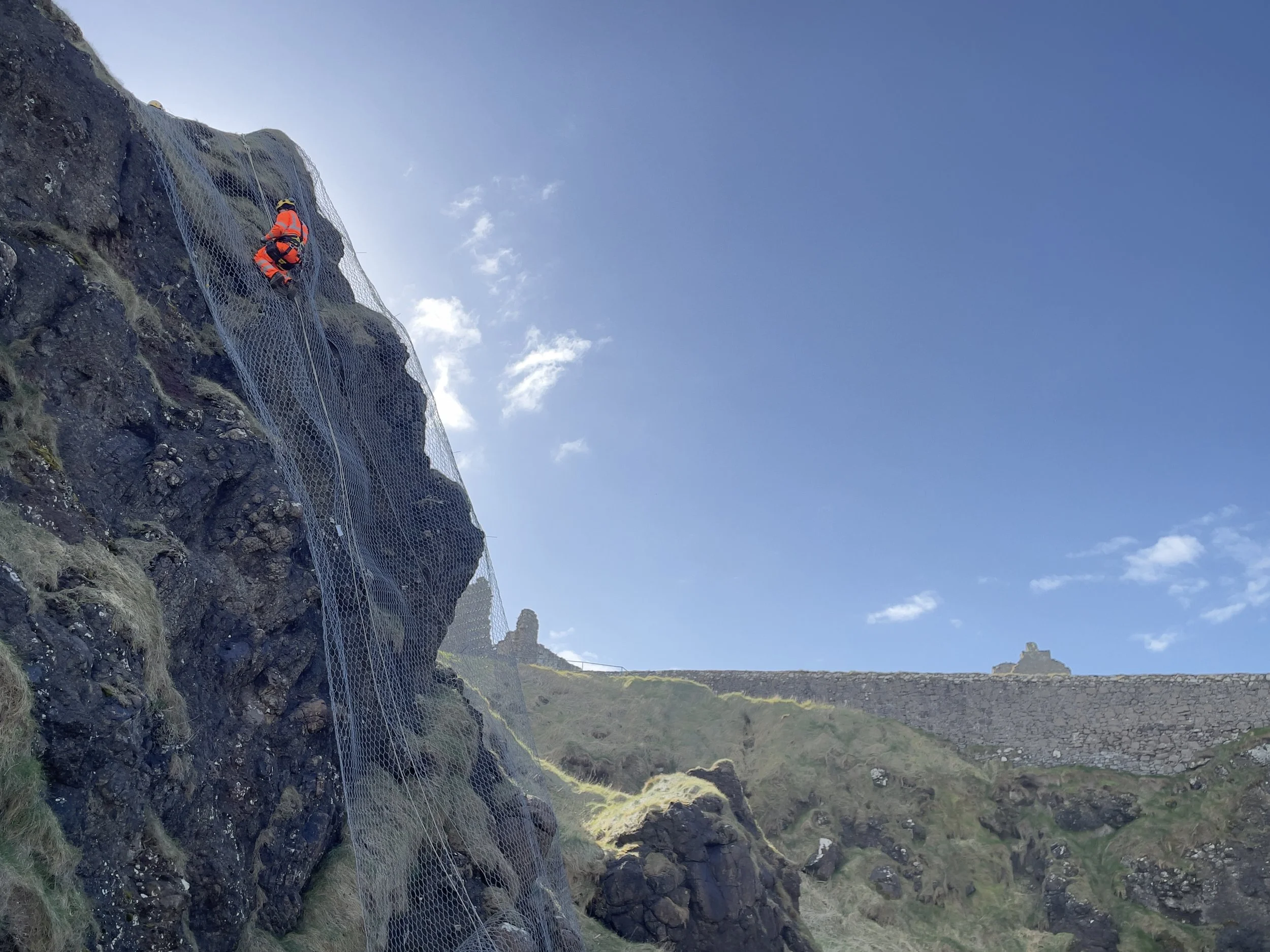 A person in orange safety gear climbing a rocky cliff with a metal safety net, under a clear blue sky, with an ancient stone wall and ruins in the background.