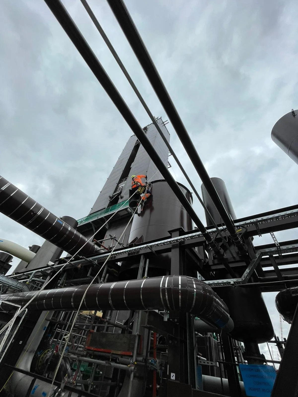 Worker in safety gear cleaning or inspecting a large industrial structure on an oil or gas platform, with pipes and metal framework, under a cloudy sky.
