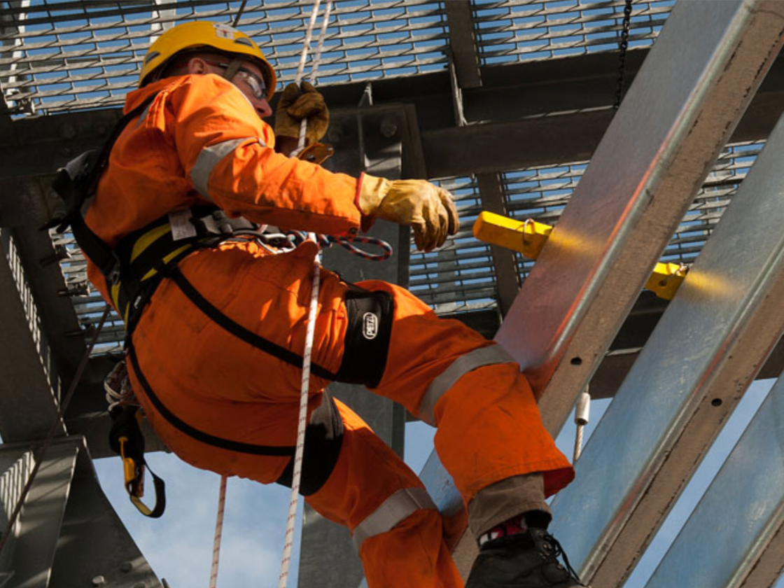 Worker in orange safety gear cleaning or inspecting a large industrial structure at an oil or chemical refinery with pipes and machinery.