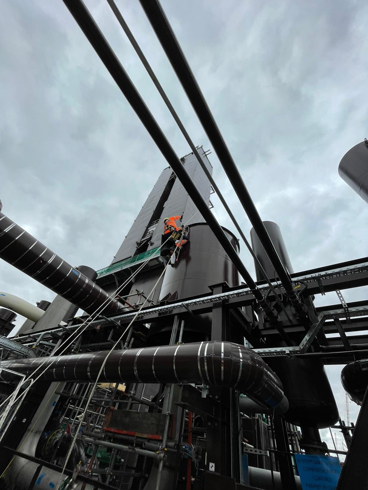 Worker in orange safety gear cleaning or inspecting a large industrial structure at an oil or chemical refinery with pipes and machinery.