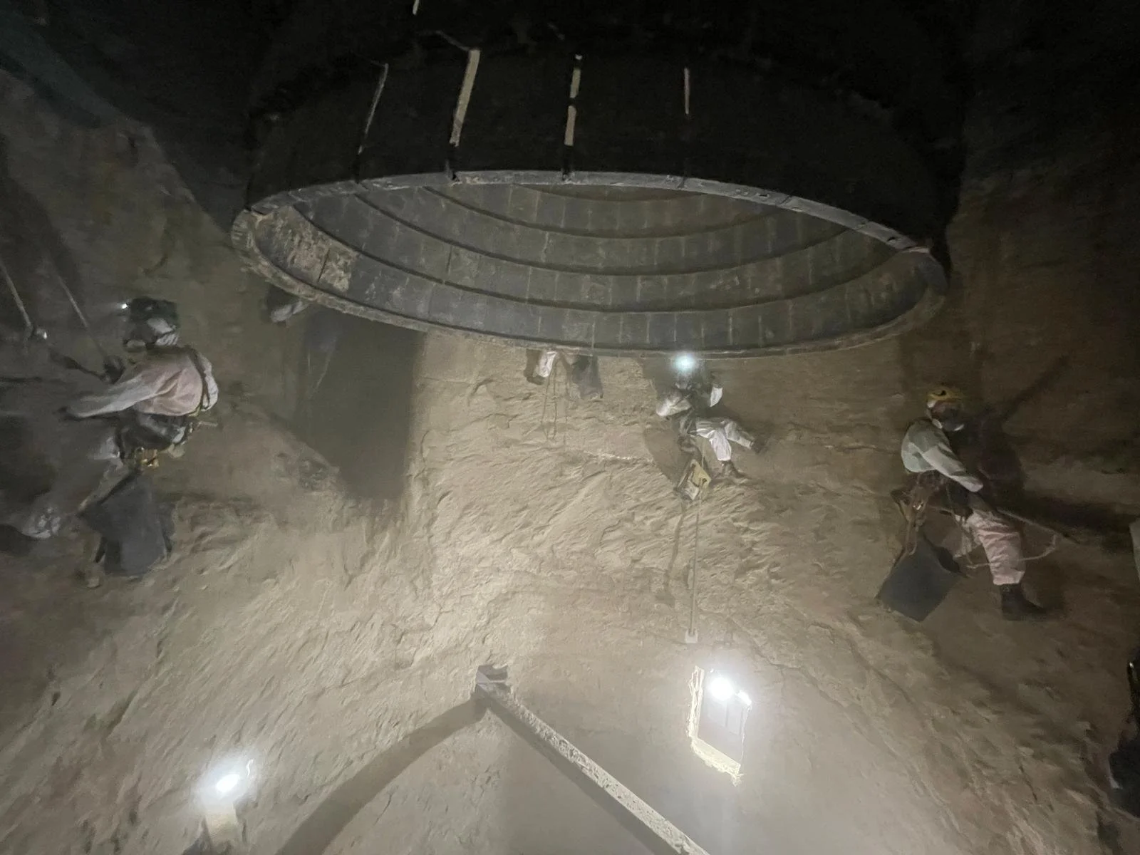 Workers underground in a mine, wearing helmets and safety gear, operating a large piece of mining equipment.