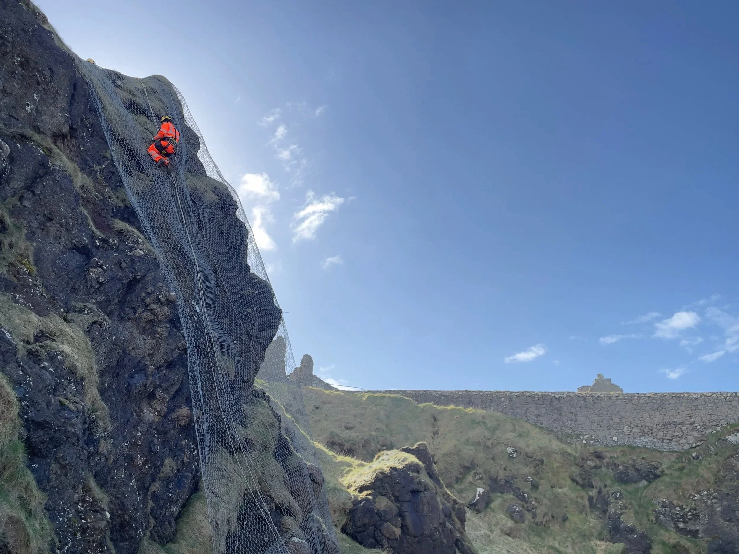 A person wearing an orange safety vest and helmet is climbing a steep, rocky cliff using a rope and mesh safety netting.
