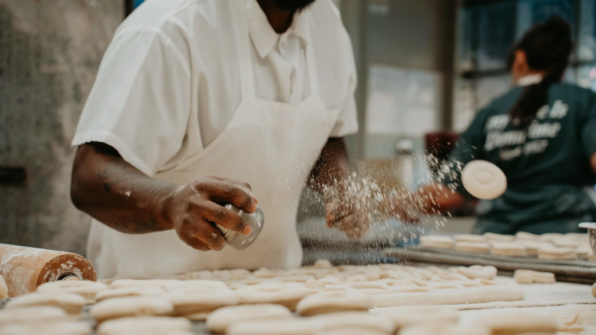 A baker in a white shirt and apron sprinkles flour on a large tray of dough pieces in a bakery kitchen, with another person in the background.