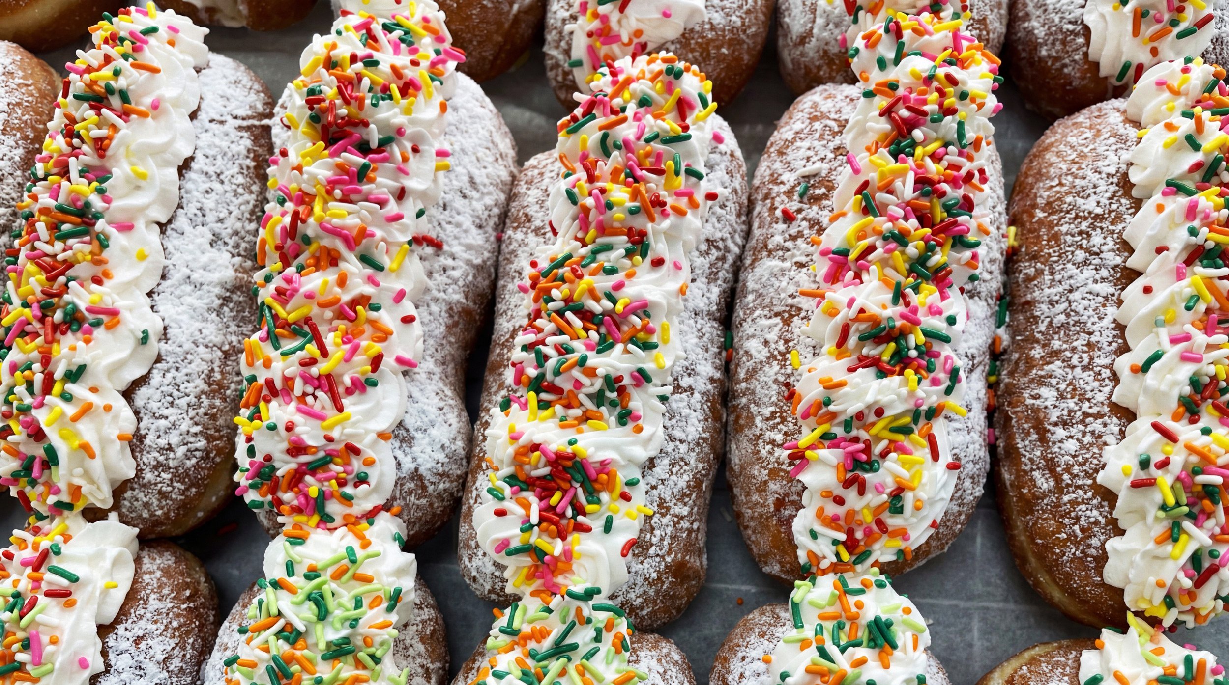 Eclairs topped with whipped cream and colorful rainbow sprinkles, dusted with powdered sugar.