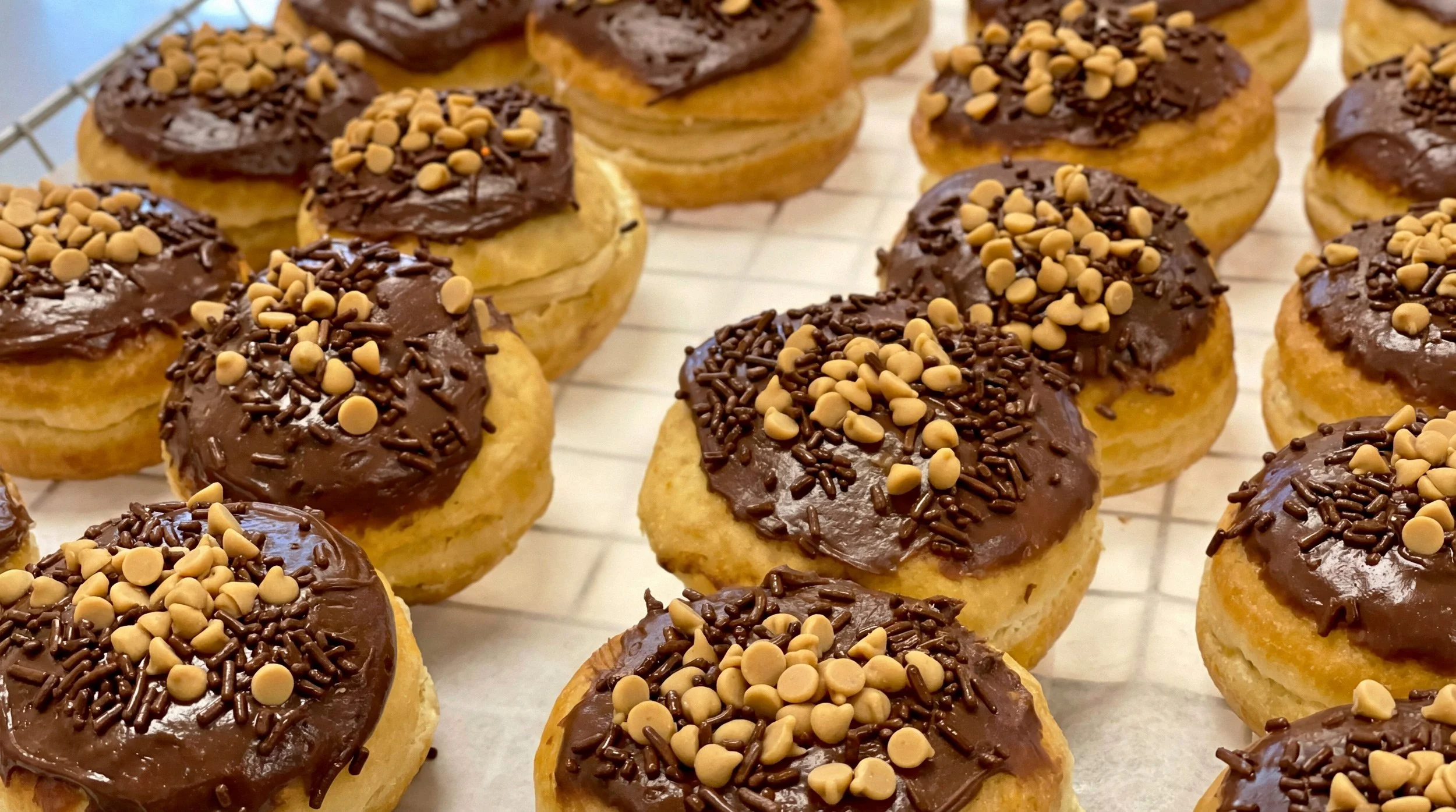 A tray of glazed donuts topped with chocolate frosting, chocolate sprinkles, and beige-colored round candy pieces.