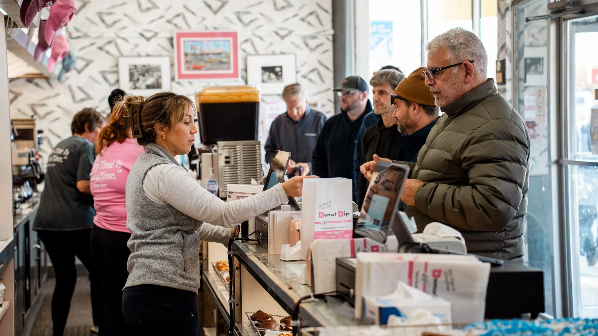 Customers ordering at the counter in a busy donut shop with a worker taking orders.
