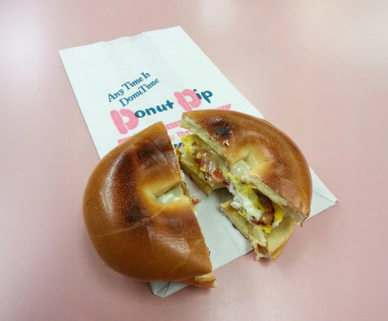 A partially eaten breakfast sandwich with egg, cheese, and bacon in a donut bun, placed on a pink surface with a paper that says 'Any Time Is Donut Time' and 'Donut Dip'.