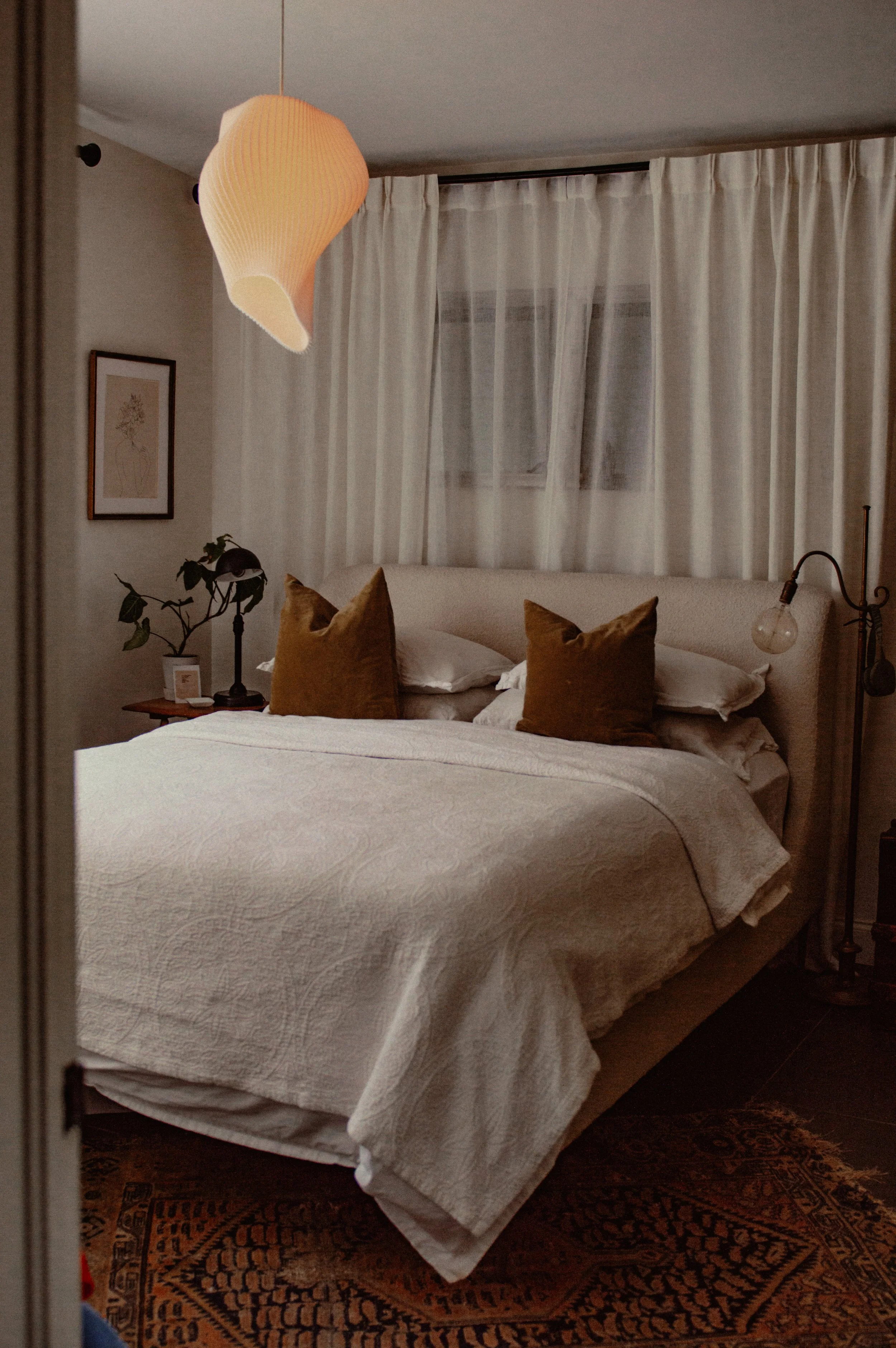 A cozy bedroom with a bed, brown pillows, and white bedding, illuminated by warm lighting, with a bedside table, lamp, and plant in the corner.