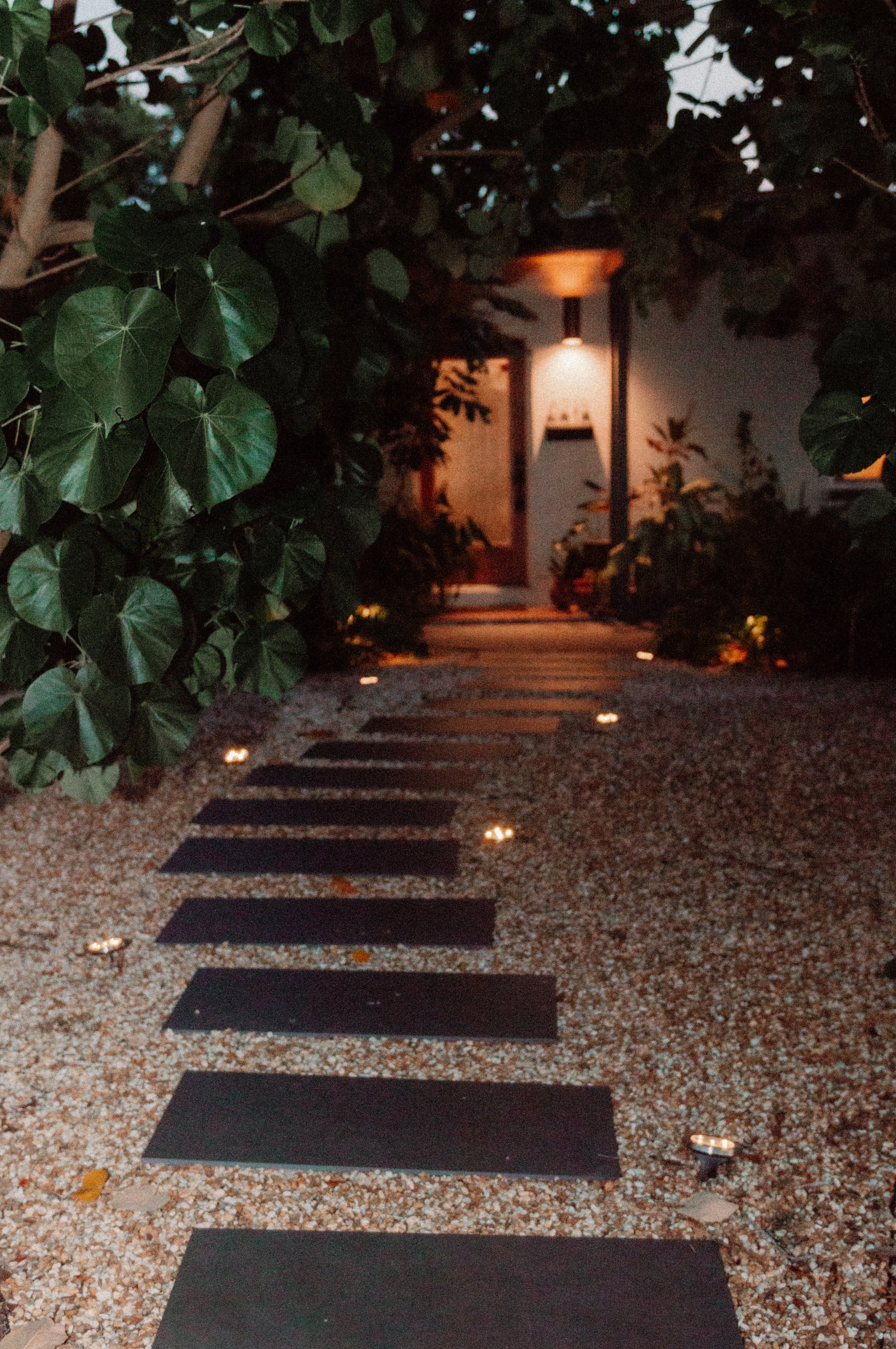 A pathway with black rectangular stepping stones illuminated by small ground-level lights, leading to a house entrance with a warm outdoor lamp, surrounded by lush green bushes and trees at dusk.