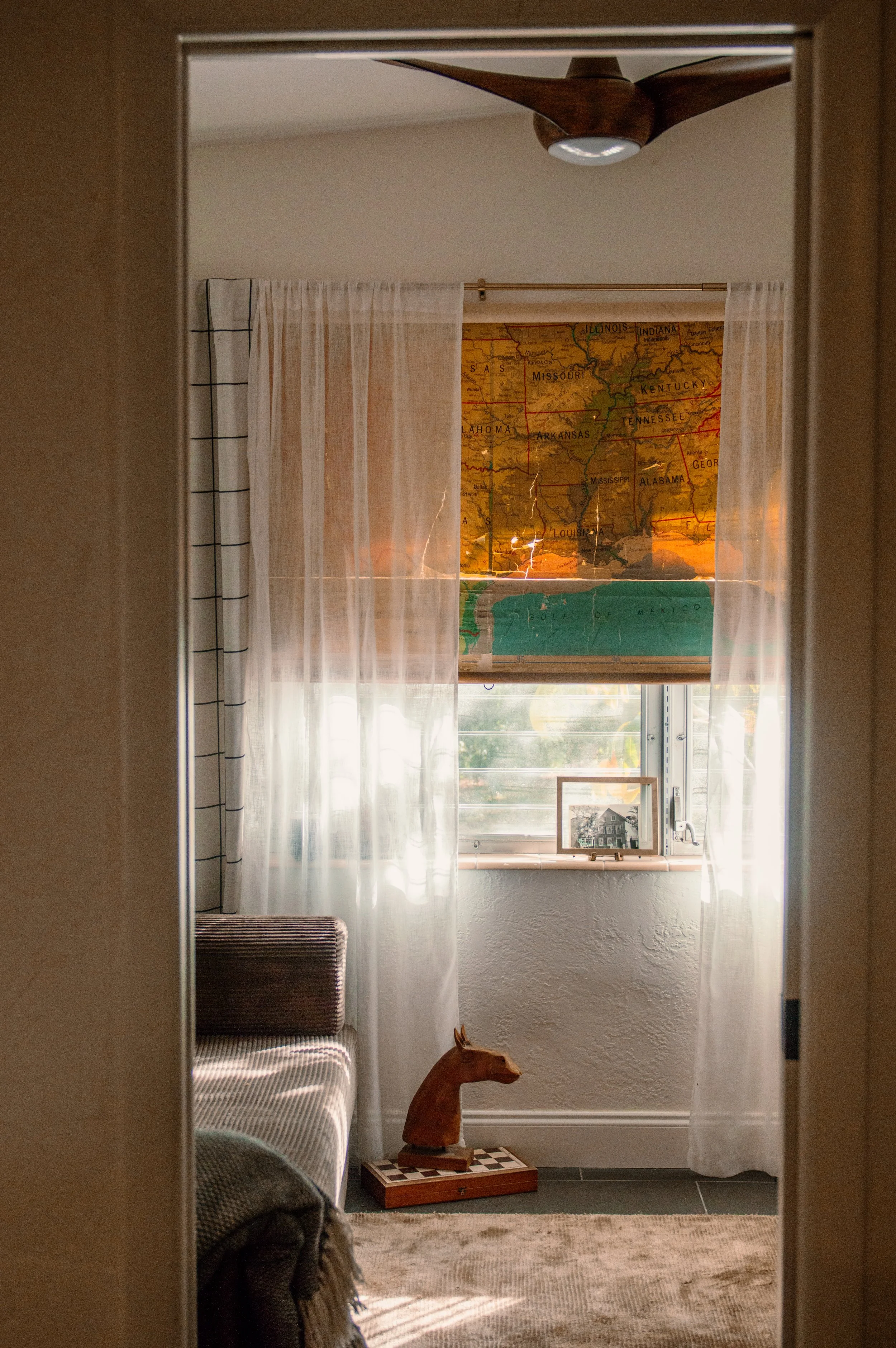 View of a sunlit room seen through an open doorway, with a wooden horse sculpture on a chessboard, a window with sheer curtains, and a wall map of the central U.S. with a ceiling fan above.