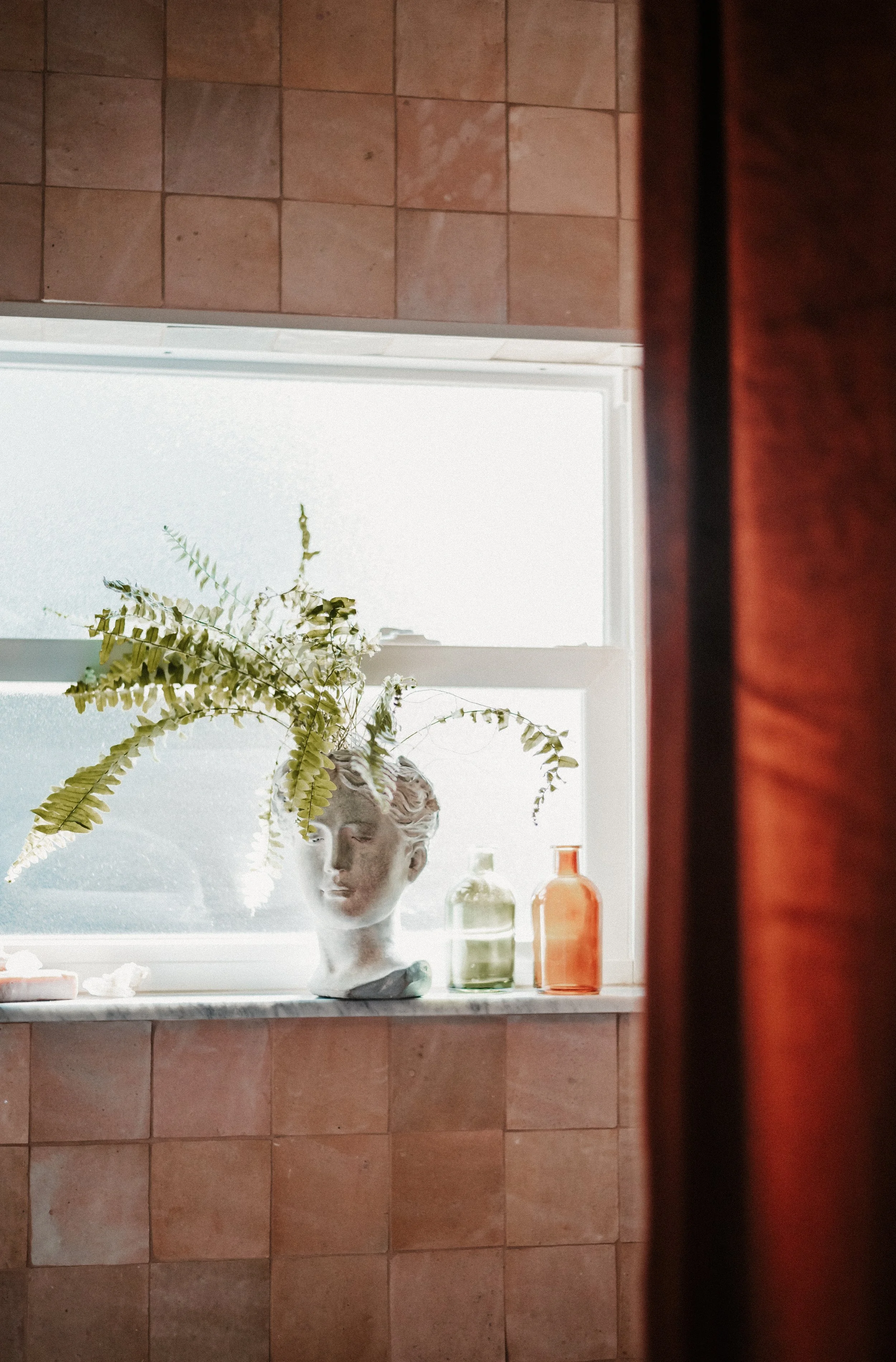 Decorative window ledge with a bust vase holding fern ferns, and two small colored bottles, one green and one amber, in a room with peach-colored tiled walls and a reddish-brown curtain.