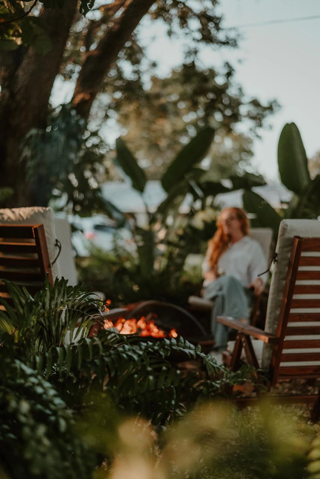 A blurred woman sitting outdoors on a wooden chair in a garden, with green foliage and trees around, during sunset or evening.