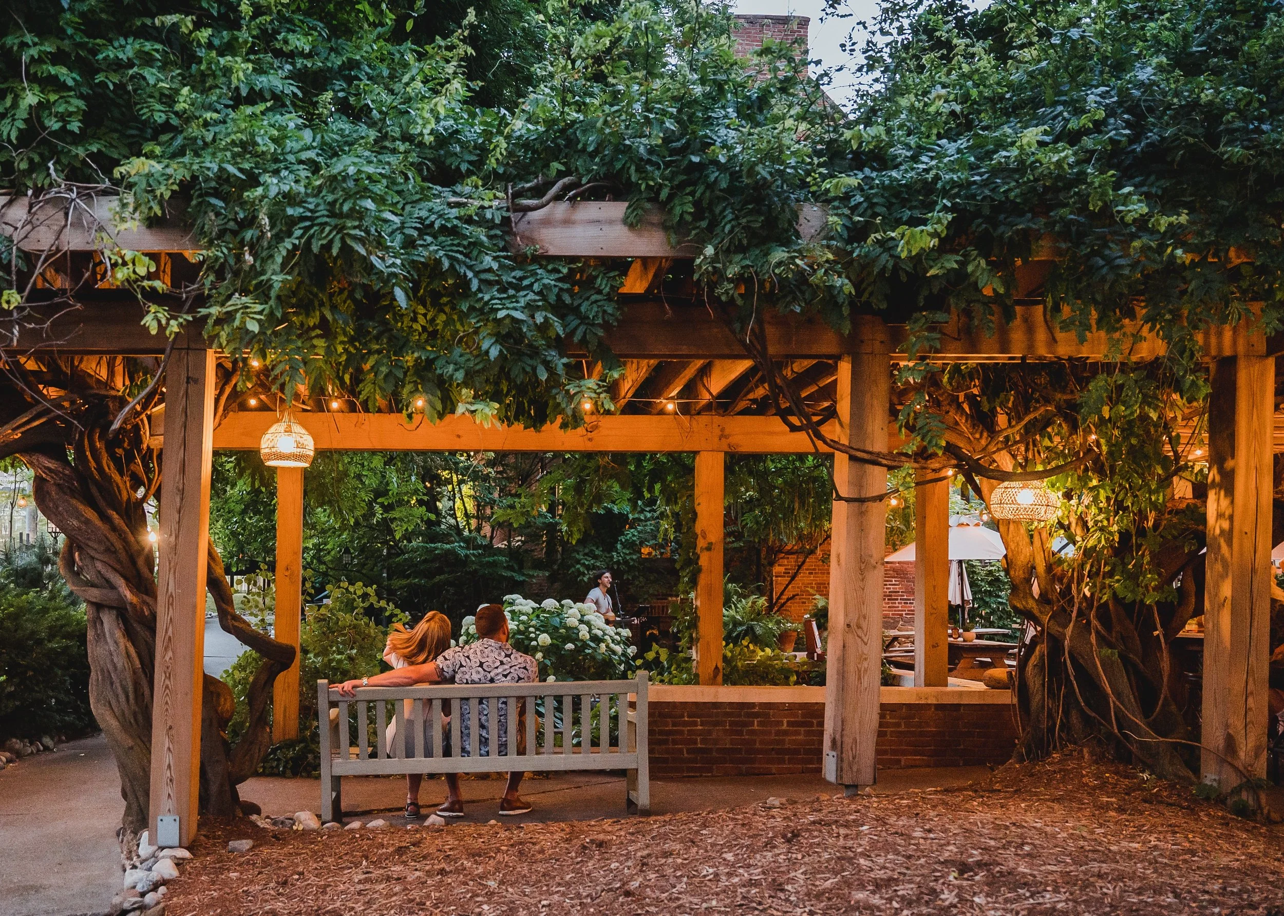 Couple sitting on a bench in a garden patio, surrounded by lush green trees and illuminated by warm outdoor lighting.