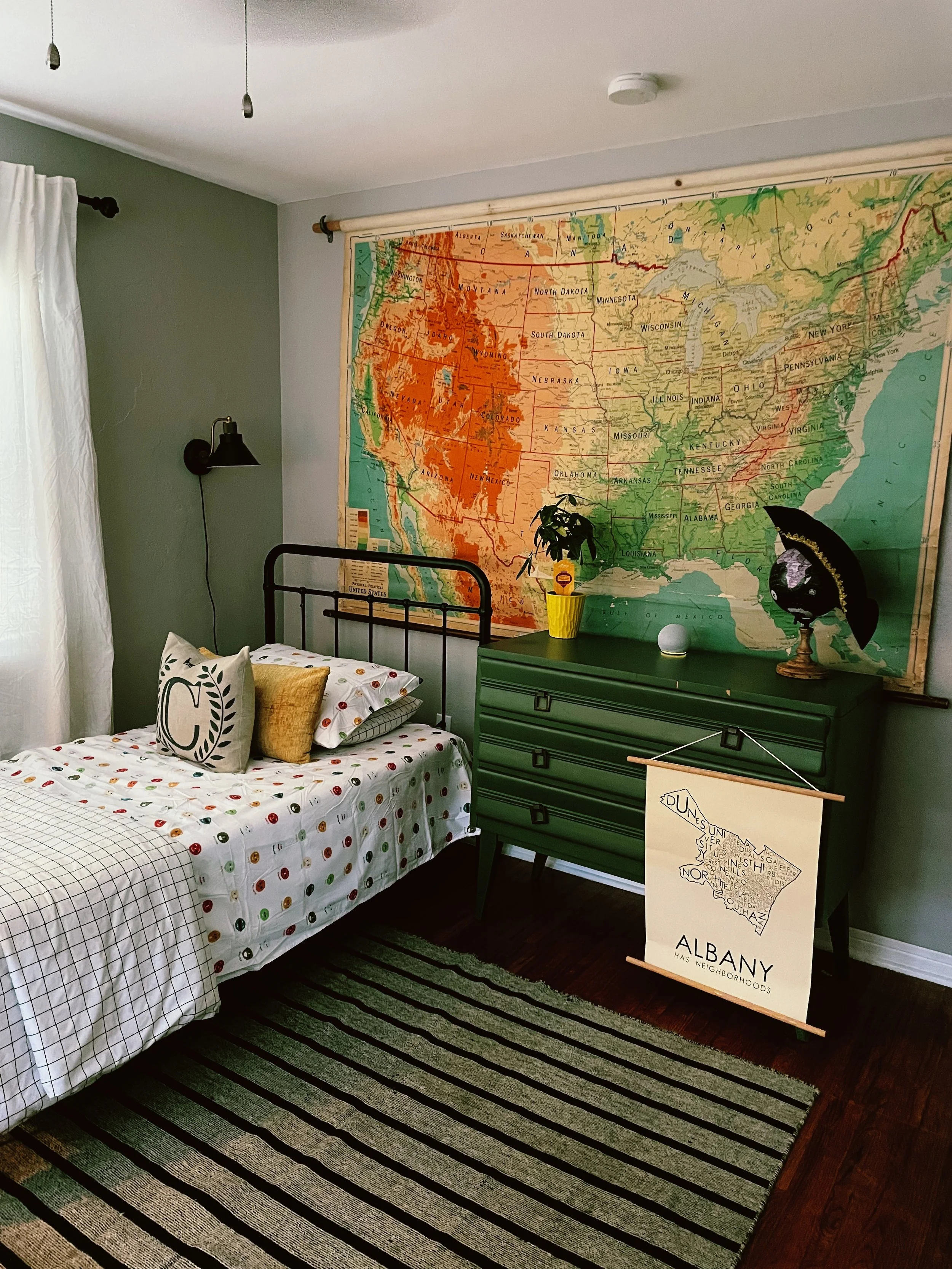 A bedroom with a twin bed, green side table, large map of the United States on the wall, and various decorative items.