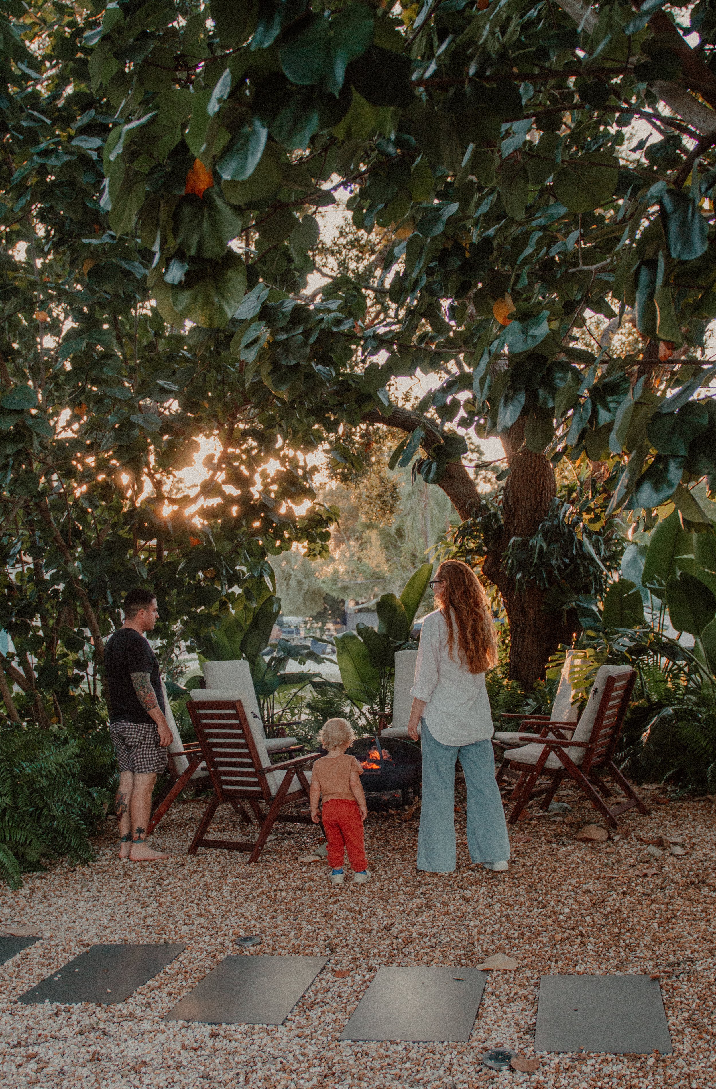 Three people — a young woman, a young man, and a small child — are gathered around a small outdoor fire pit in a lush garden setting during sunset. Two wooden chairs are placed nearby, surrounded by large leafy plants and trees.