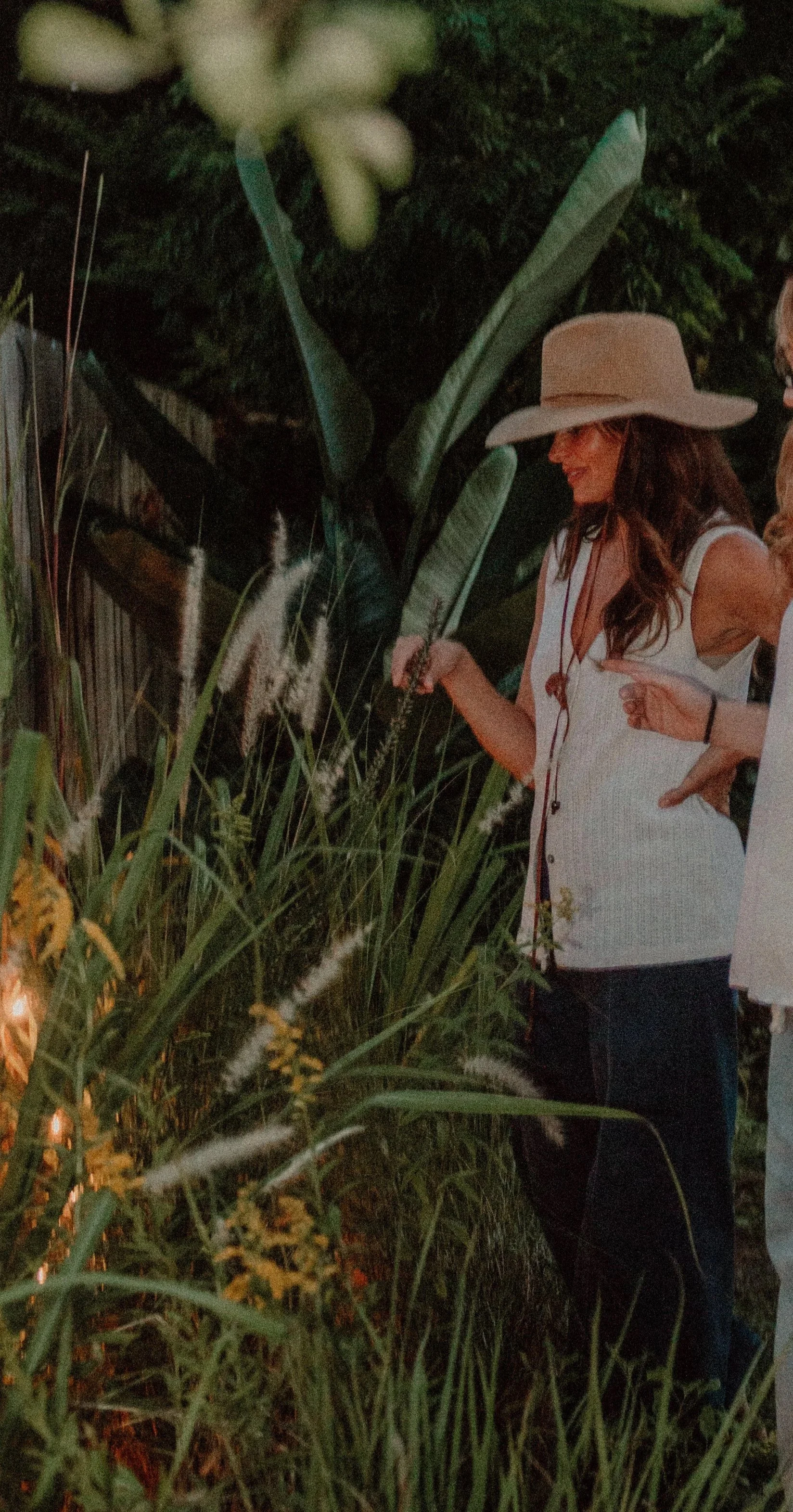 Woman wearing a large sunhat and white top, standing outdoors amidst tall grass and plants, smiling and looking at the foliage during evening or night time.