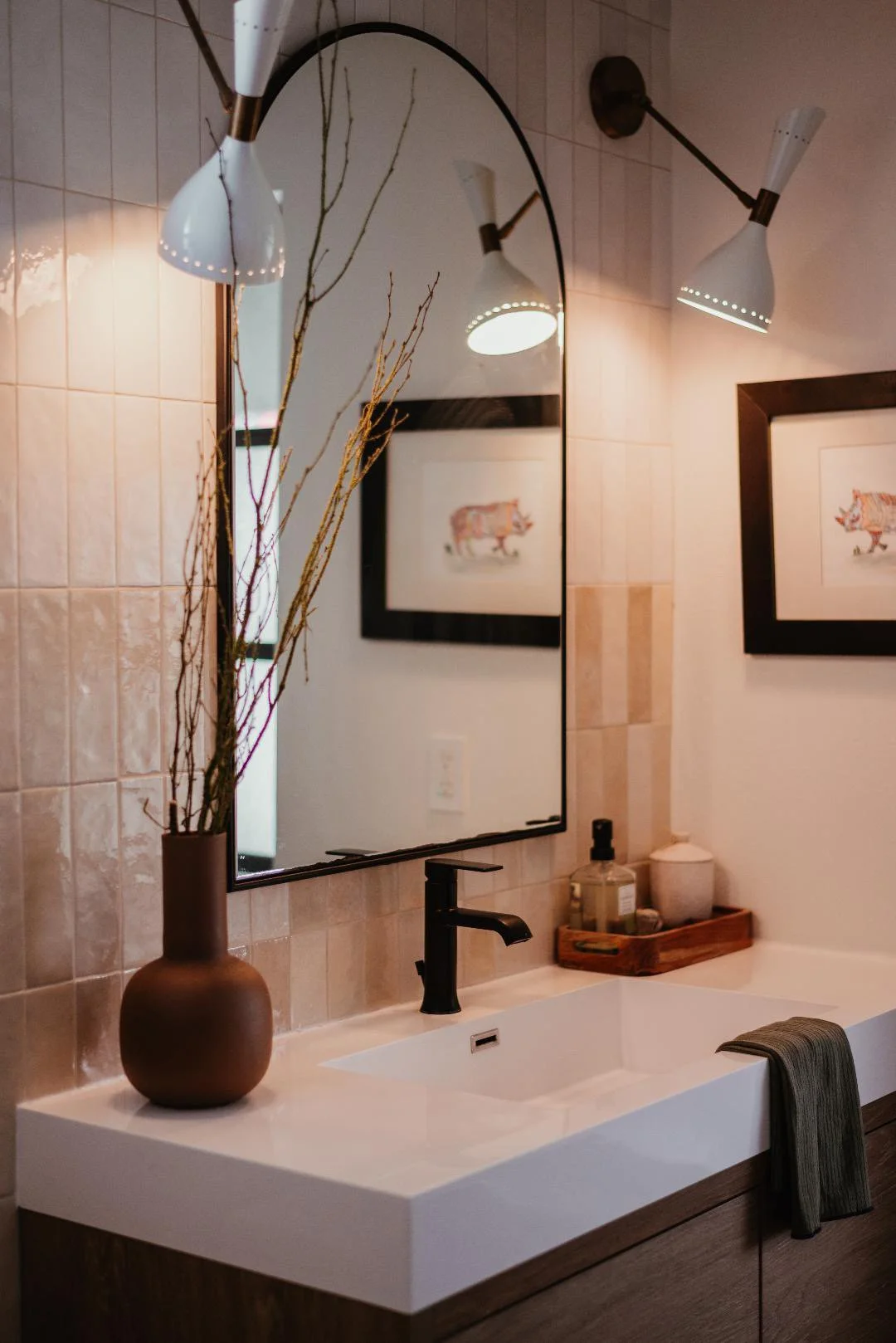 Modern bathroom with white sink, black faucet, large mirror, wall-mounted light fixtures, framed art of a pig, decorative branches in a brown vase, and small storage tray with bathroom essentials.