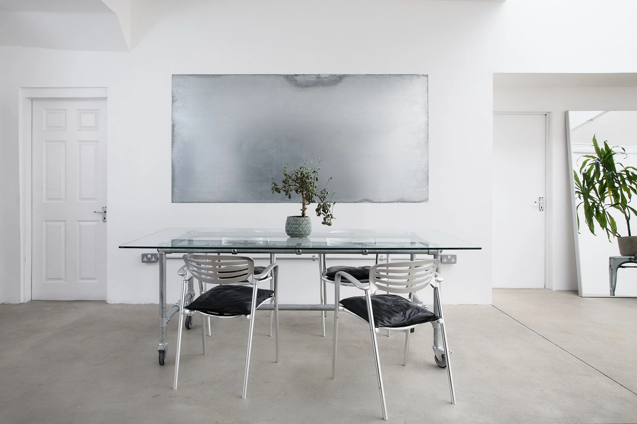 Minimalist dining room with a glass table, four white chairs with black cushions, a potted plant in the center, white walls, and a mirror leaning against the wall on the right.