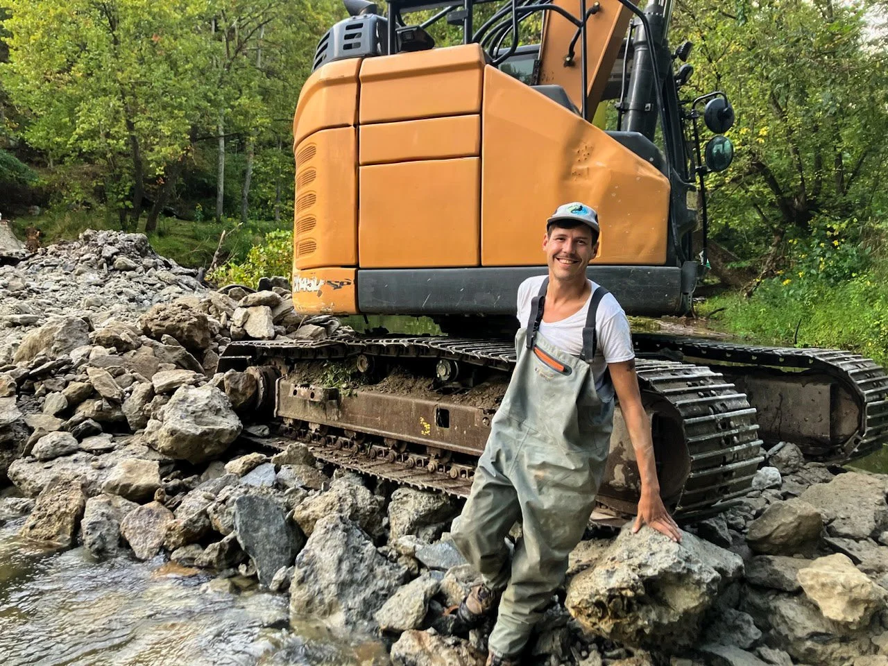 A man smiling and wearing waders standing on rocks in a stream, with a large orange excavator behind him in a forested area.