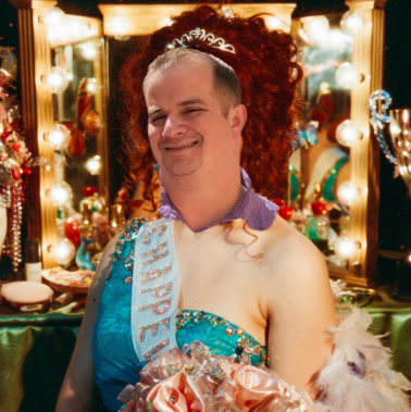 A man dressed as a princess stands in front of a decorated vanity mirror with lights, wearing a tiara, a sash, and a blue dress, smiling at the camera.