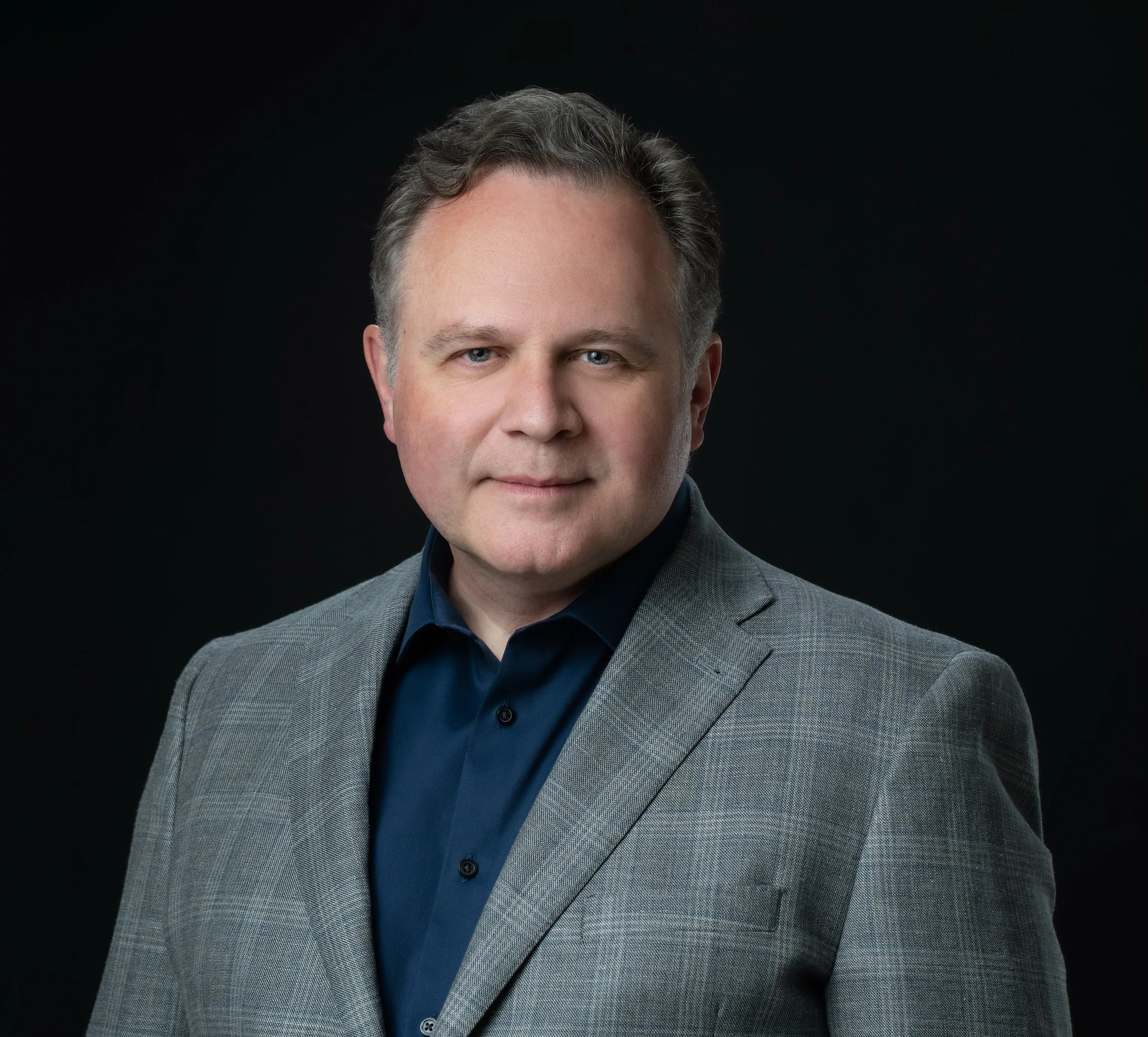 Professional portrait of a middle-aged man with gray hair, wearing a gray suit jacket and a navy blue shirt, against a black background.