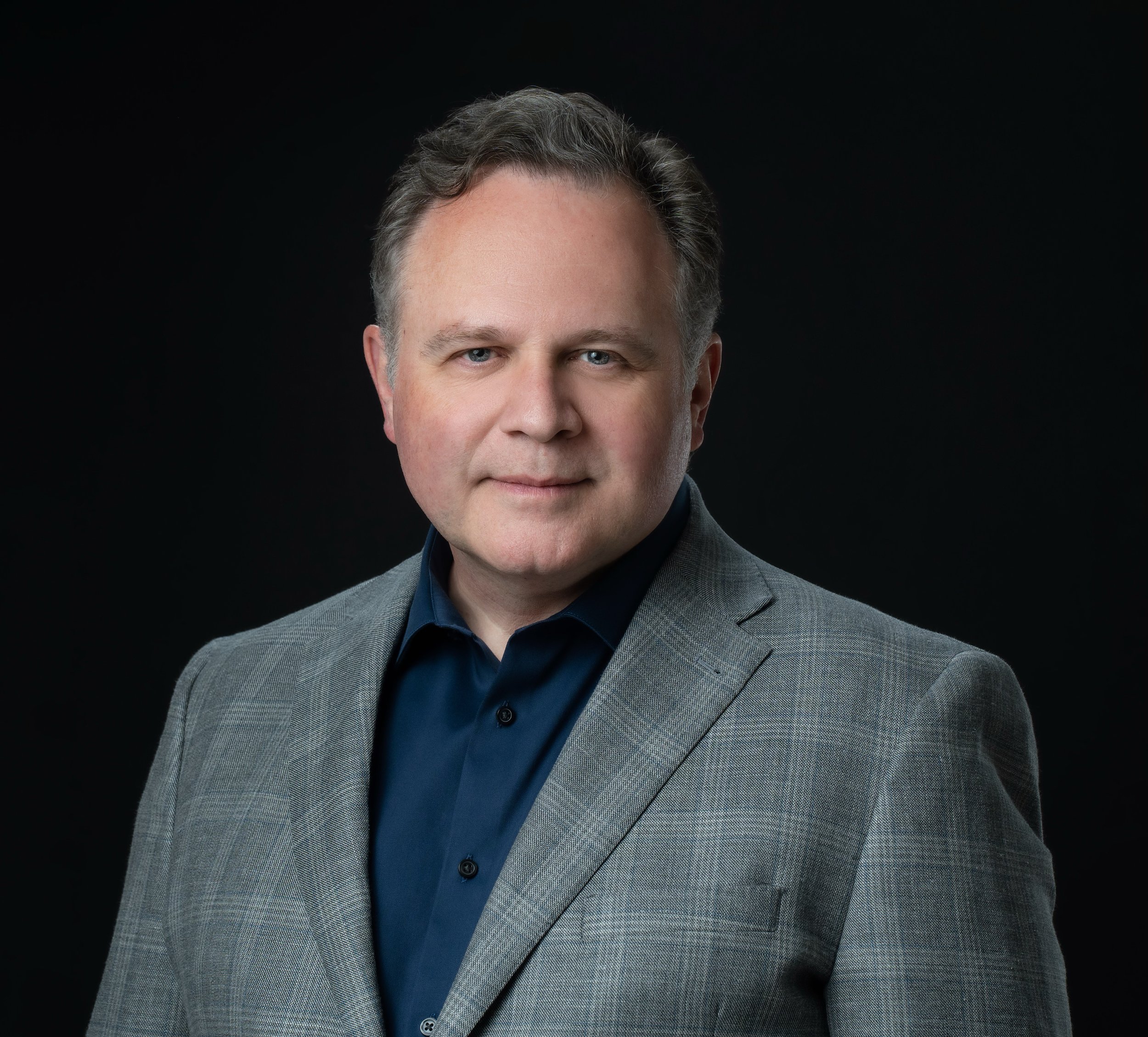 Professional headshot of a middle-aged man with short, wavy brown hair, wearing a gray checkered blazer and a dark blue dress shirt, against a black background.
