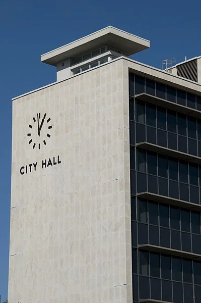 A tall modern city hall building with a clock showing 12:00, featuring a combination of beige stone and dark glass windows, with a clear blue sky in the background.