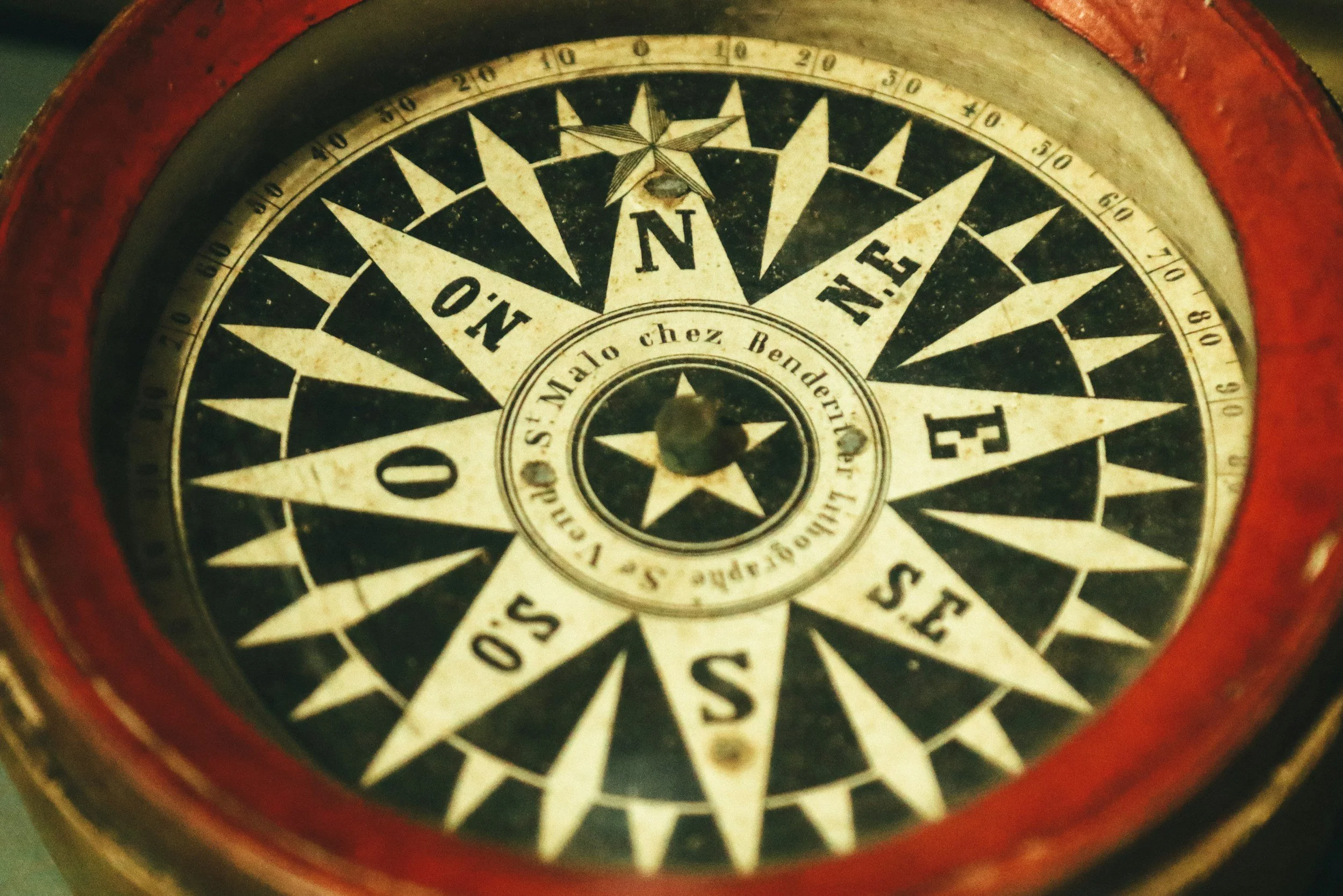 Close-up of a vintage compass with a red border, black and white face, and directional markers including 'N', 'E', 'S', and 'O', with the words 'Chez Bender' and 'Vittorio S. Malò' on it.