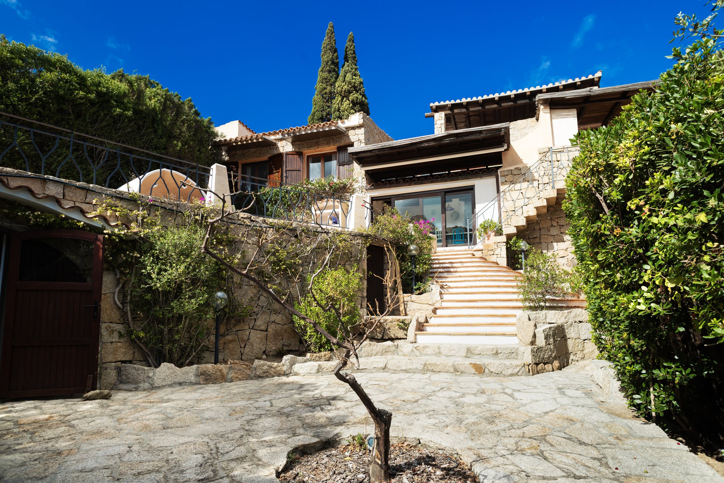 Casa di pietra con scale e giardino, circondata da alberi e cespugli, sotto un cielo blu