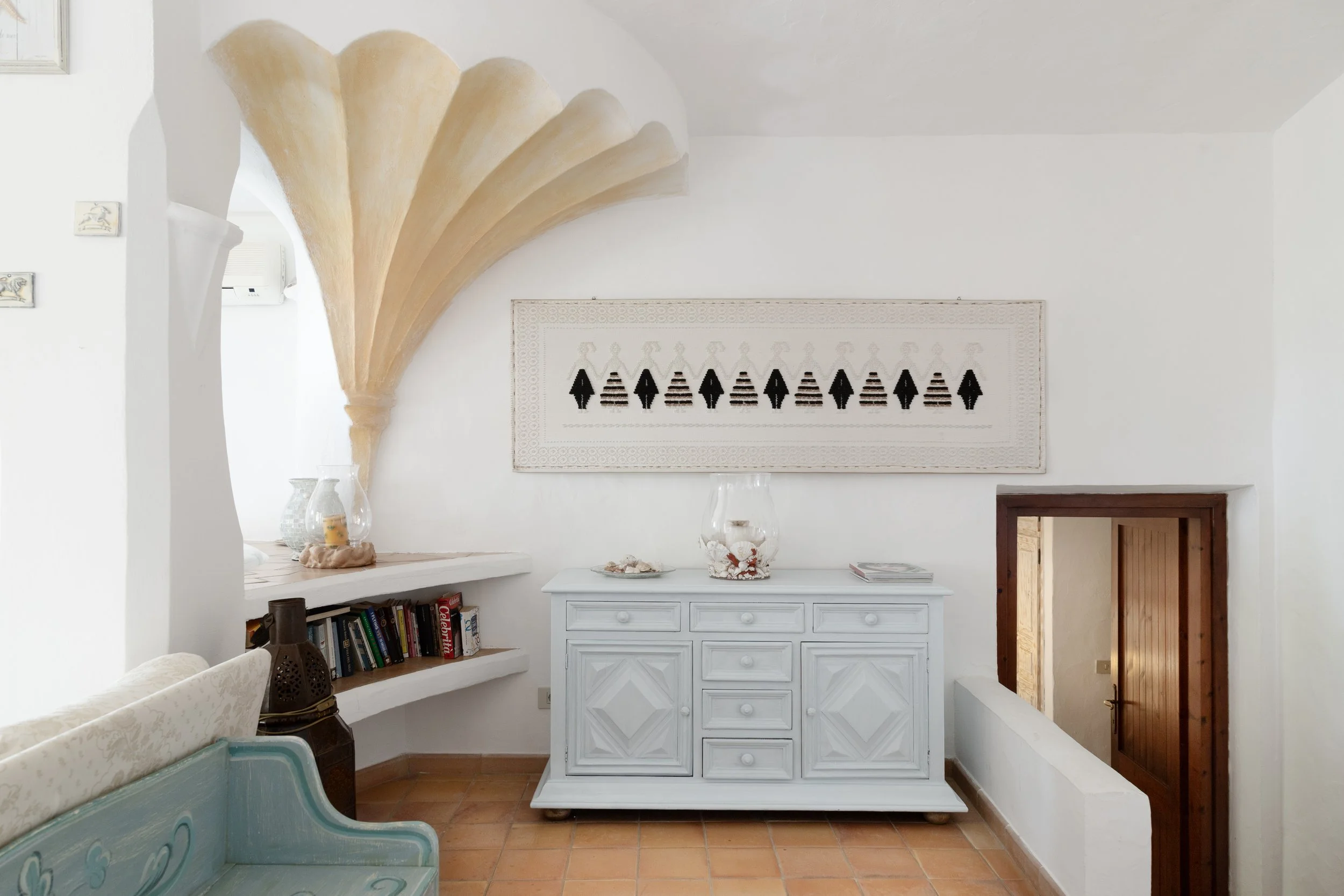 Interno di una stanza con pareti bianche, decorazioni e mobili di stile mediterraneo. Sopra una credenza bianca decorata con conchiglie, al centro di una parete, un quadro ricamato con figure di donne e uomini stilizzate, e un'arcata decorativa in al