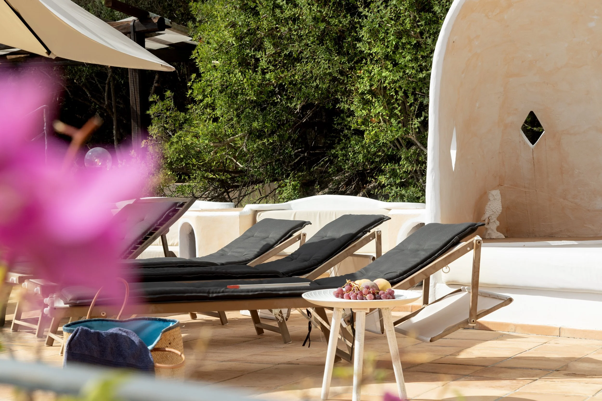 Tre lettini neri con cuscini sulla terrazza di una casa bianca con pareti di colori caldi, circondati da piante verdi.