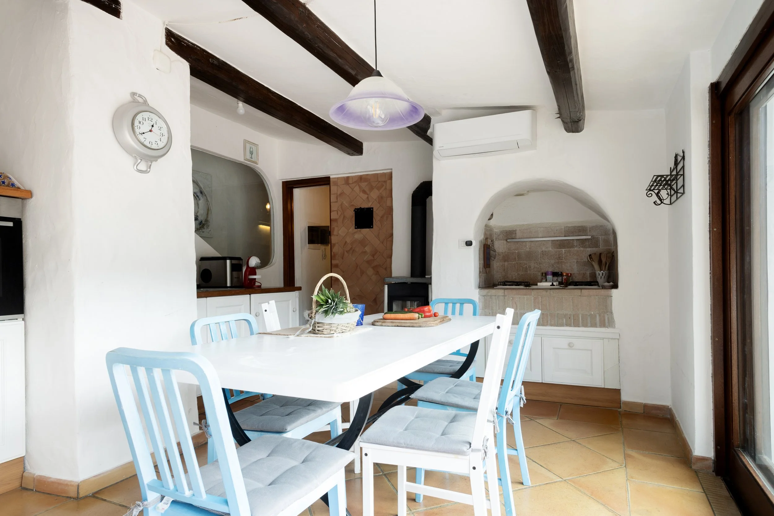 Interno di una cucina con pareti bianche, tavolo bianco e sedie blu e bianche, camino in muratura, e grandi finestre con tende di tessuto, con decorazioni rustiche e moderna.