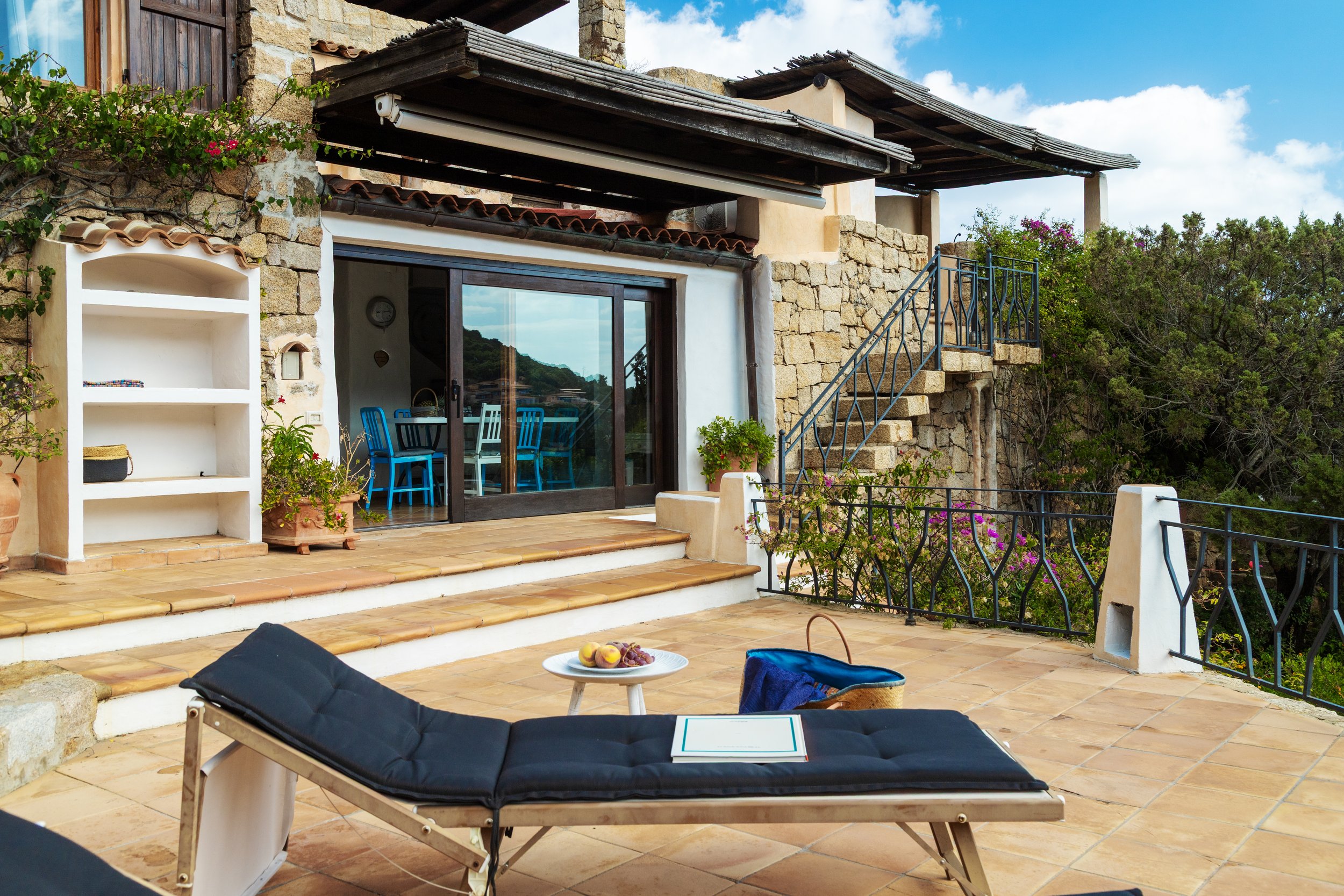 Terrazza con lettino, tavolo con frutta, vicino a casa in pietra, con piante e fiori, vista su collina e cielo azzurro con nuvole.