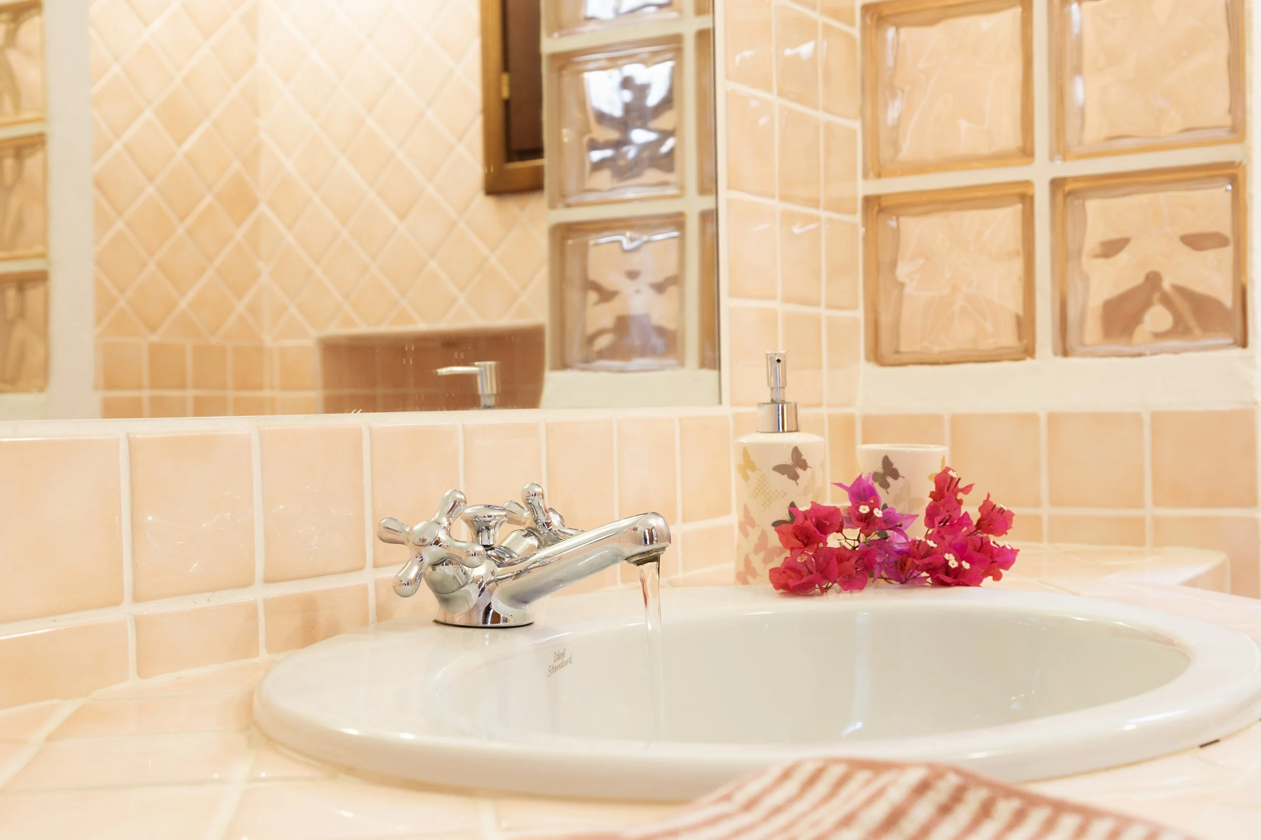 Lavandino da bagno con rubinetto e acqua corrente, dispenser di sapone e fiori rosa su un piano di piastrelle color pesca, con uno specchio e pareti di mattonelle uguali sullo sfondo.