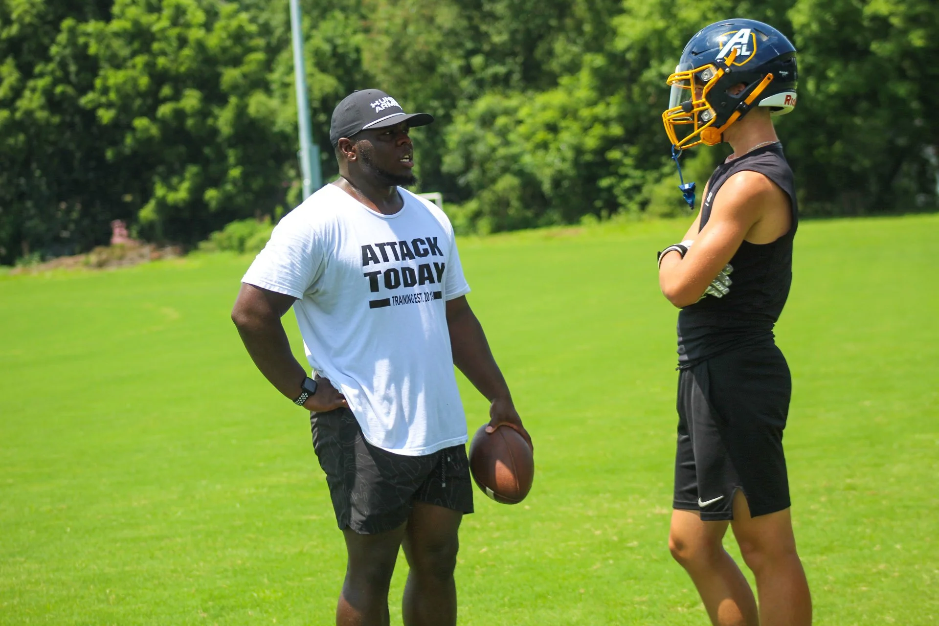 Two young men practicing football on a lush green field with trees in the background. One man is holding a football, and the other is wearing a football helmet and black athletic clothing.