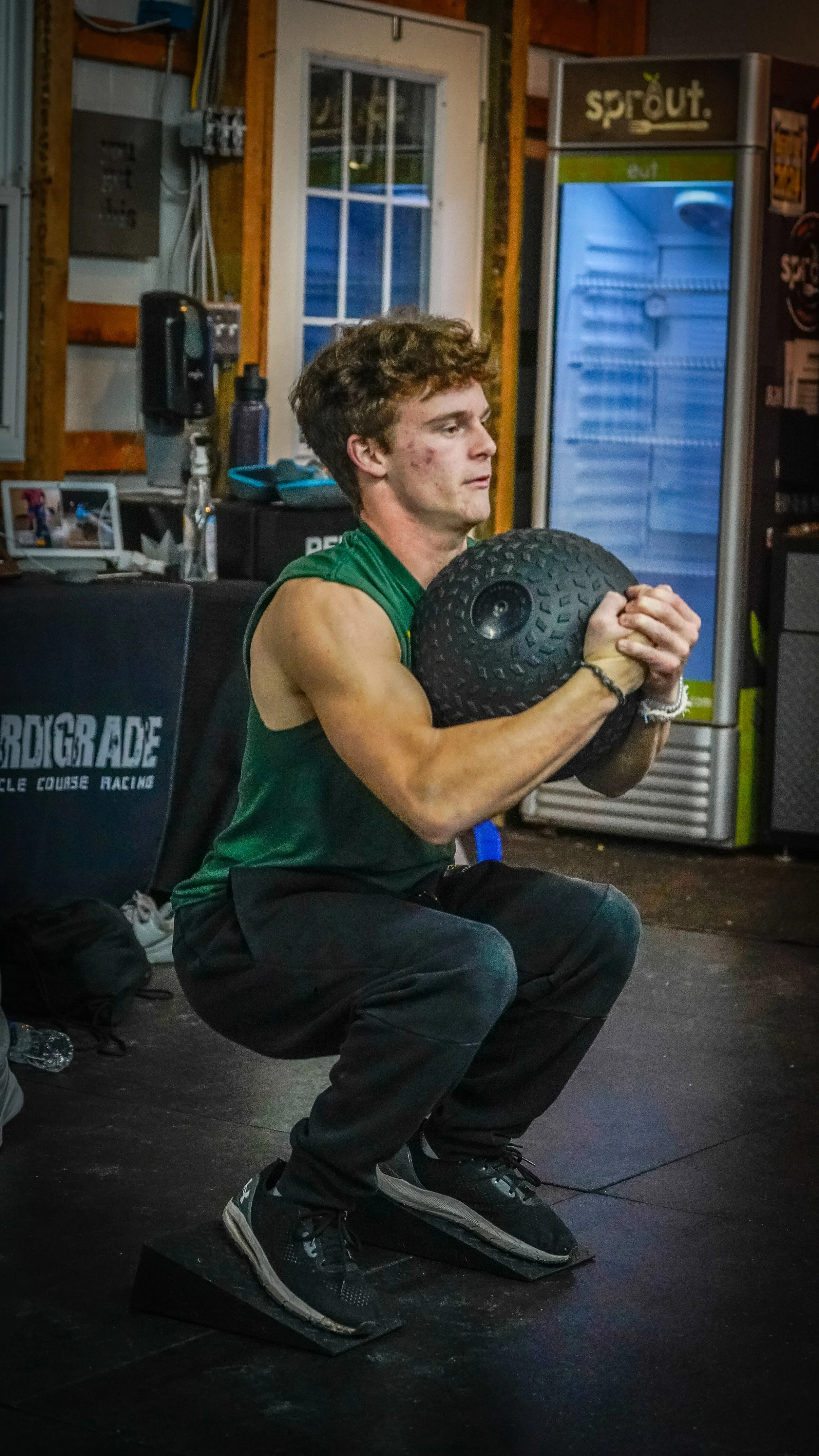 A young man in athletic clothing squatting on a box in a gym, holding a medicine ball close to his chest during a workout in a gym setting.
