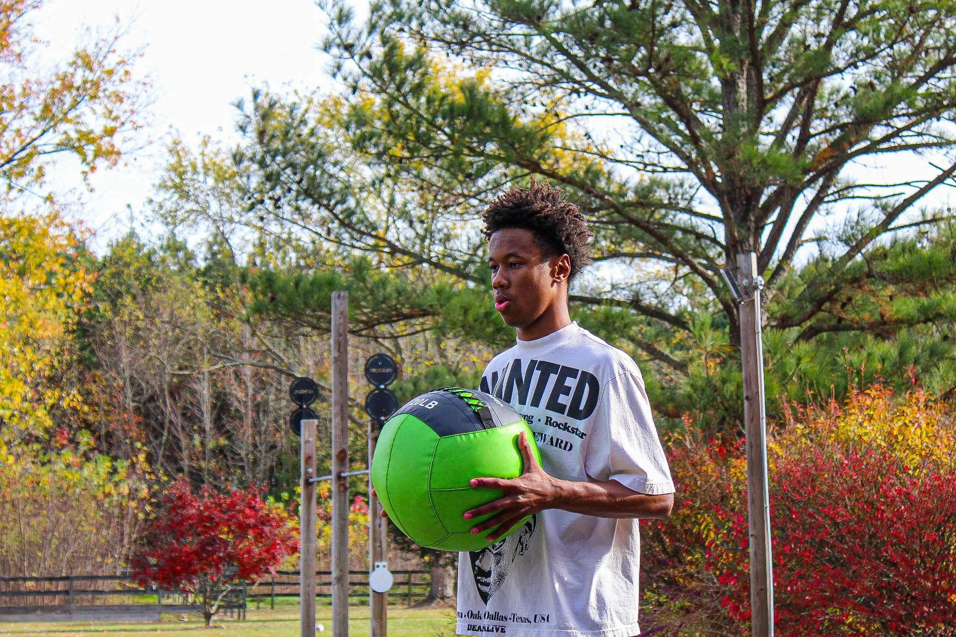 A young man holding a green medicine ball outdoors in a park with colorful autumn trees.