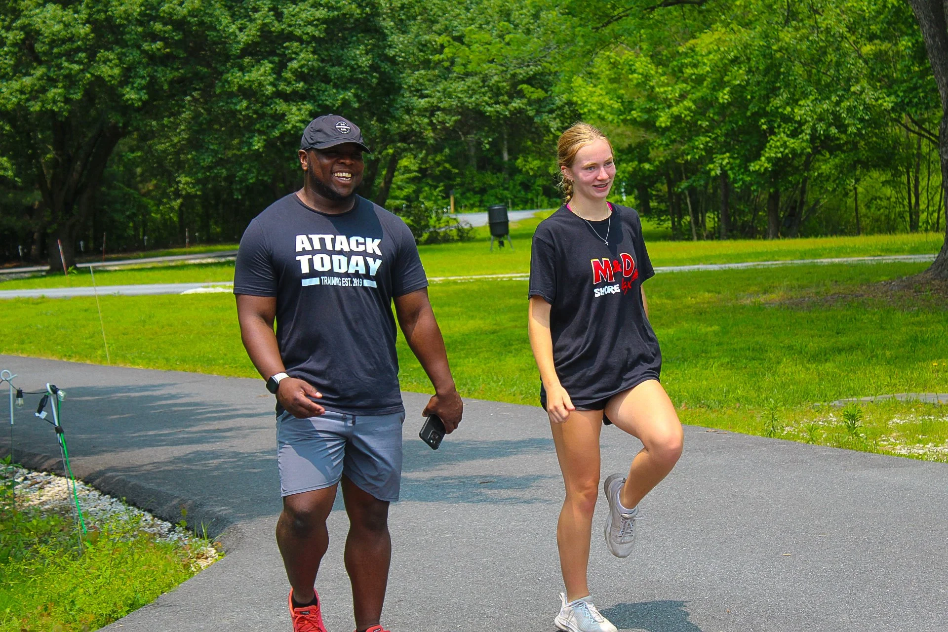 Two people, a man and a woman, jogging outdoors on a paved path surrounded by green grass and trees. The man is wearing a black cap, dark T-shirt with the text "ATTACK TODAY," and gray shorts. The woman has on a black T-shirt with some red and white 