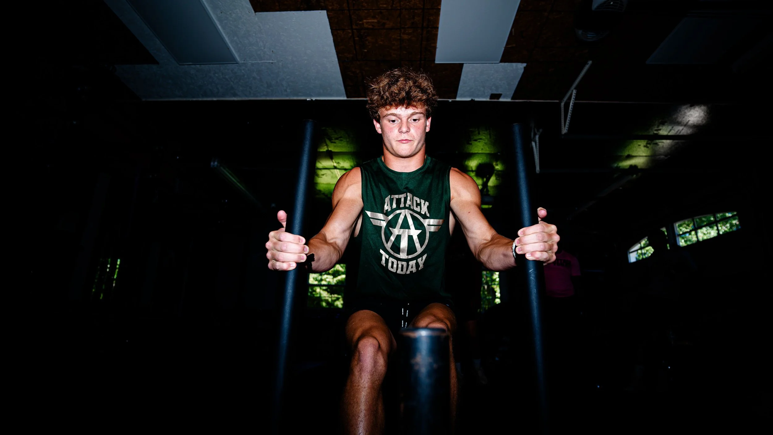 Young man with curly hair wearing a sleeveless green shirt in a gym using a rowing machine.
