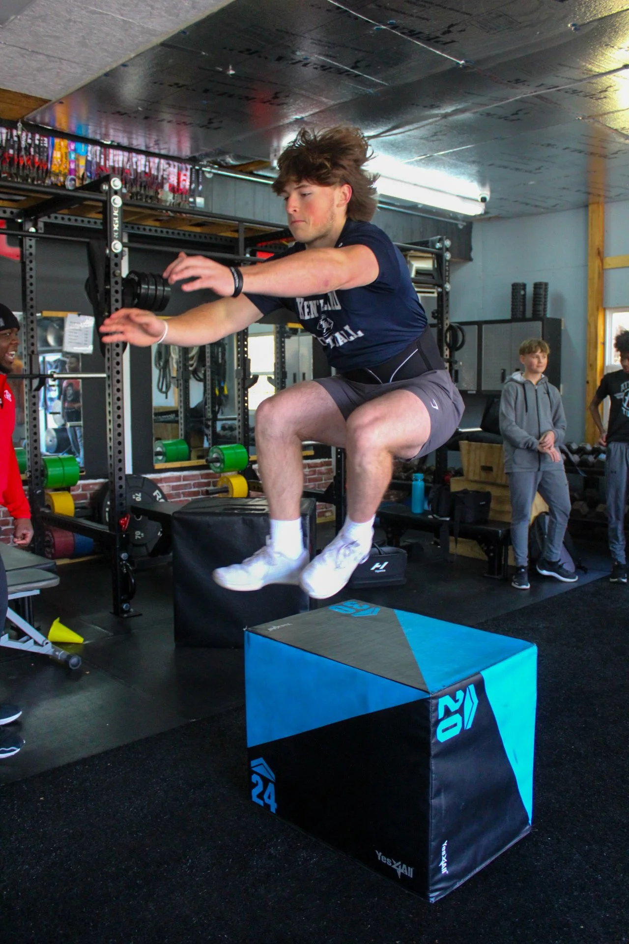 Young man jumping onto a blue and black plyometric box in a gym, with gym equipment and other people in the background.