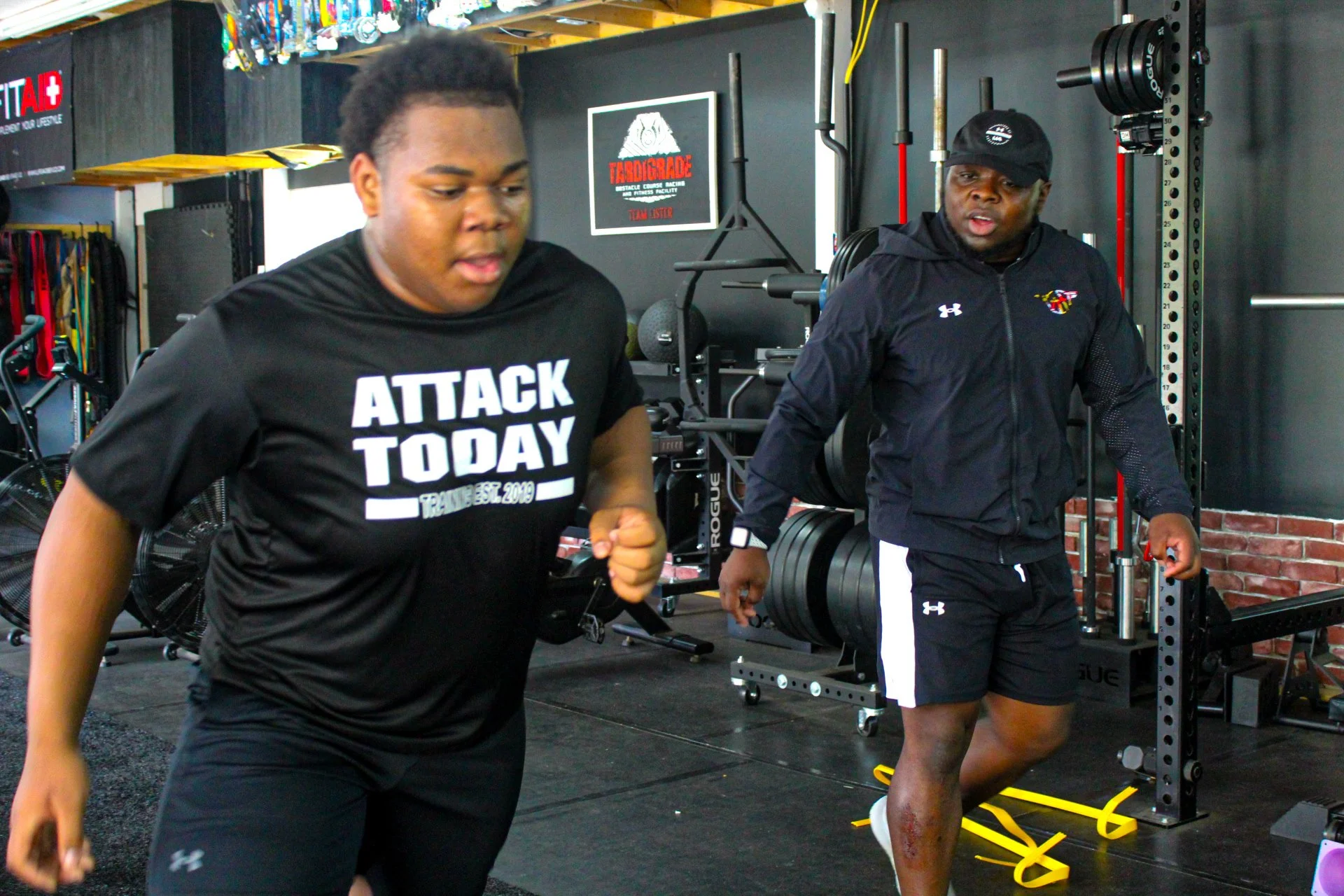 One person working out in a gym. The man in the front wears a black t-shirt with 'ATTACK TODAY' printed on it, and the man behind wears a black jacket and black shorts, standing near exercise equipment.