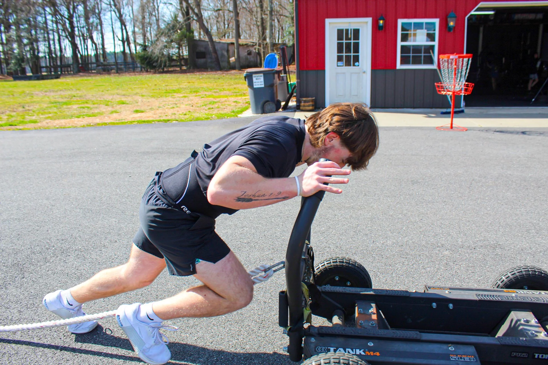 Young man in black shorts and t-shirt pulls a weighted sled on a paved driveway in front of a red and gray shed, with a basketball hoop and trees in the background.