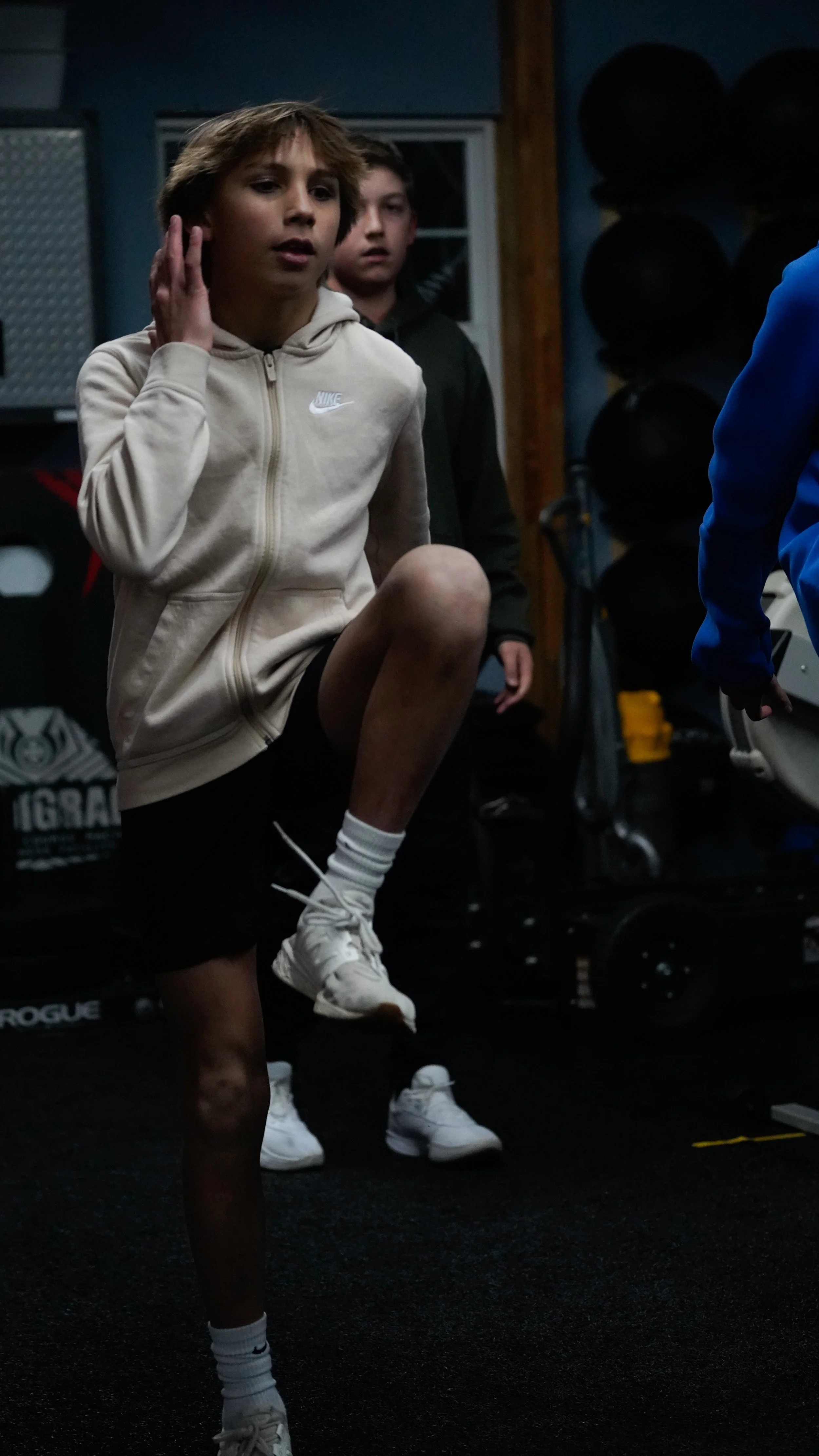 A young boy lifting his knee high during a workout at a gym with two other boys watching, gym equipment visible in the background.