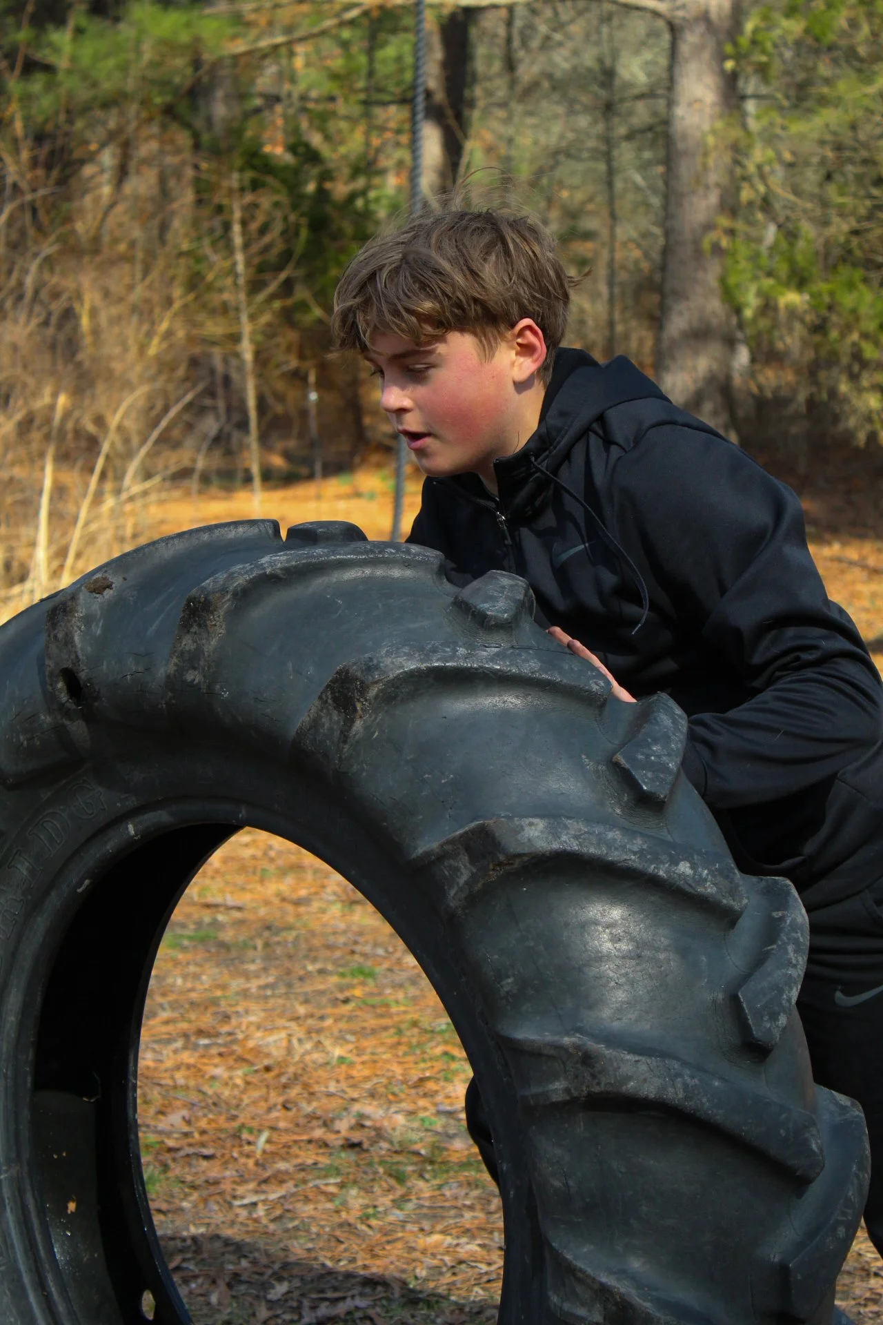 A young boy in a black jacket is pushing a large tractor tire outdoors on a leaf-covered ground with trees in the background.