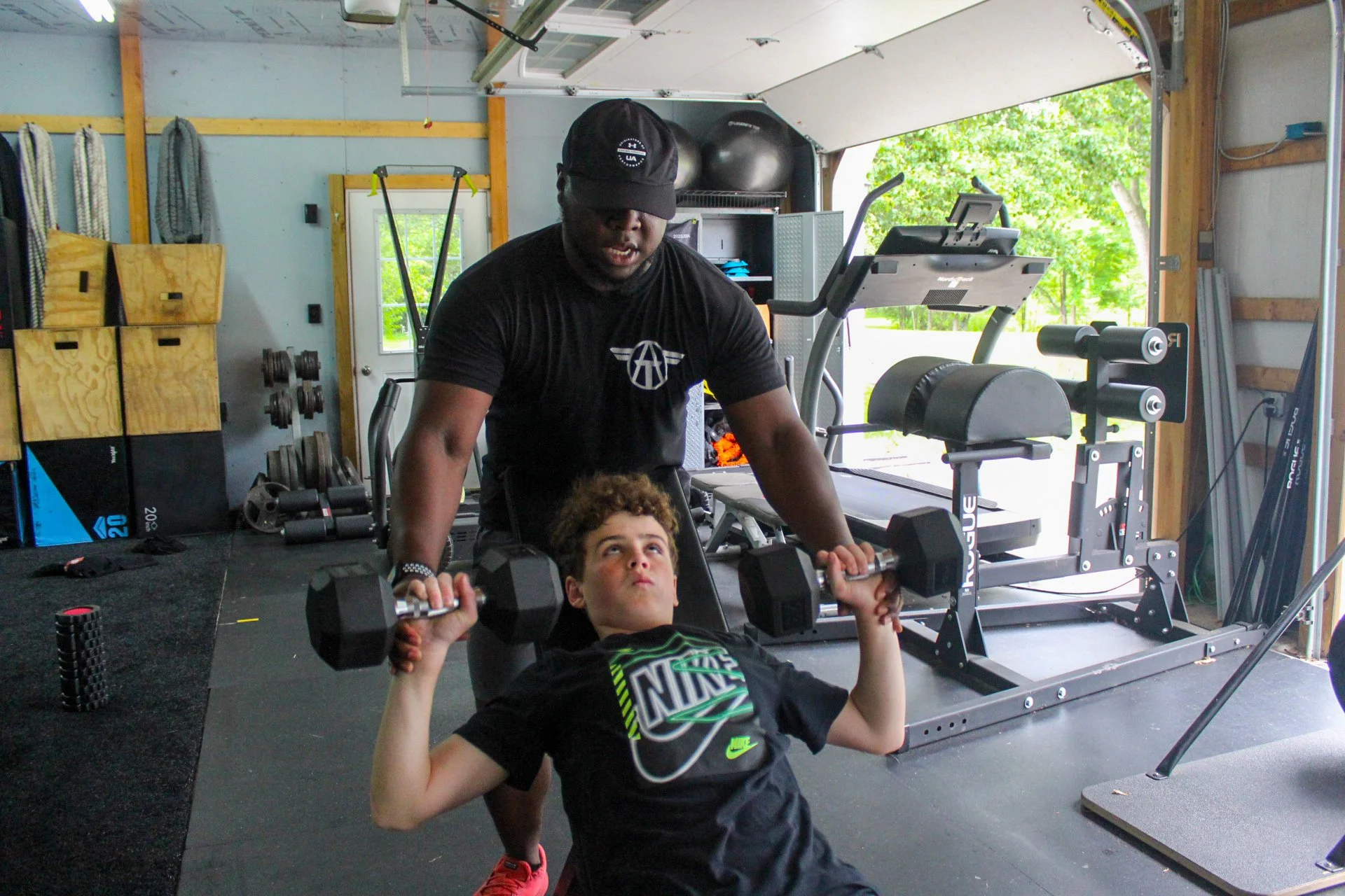 A young boy is doing a bench press exercise with dumbbells while a trainer, spots him in a gym garage. The boy is lying on a bench, lifting a pair of dumbbells, and looking upward. The gym has exercise equipment, weights, and a treadmill, with a brig