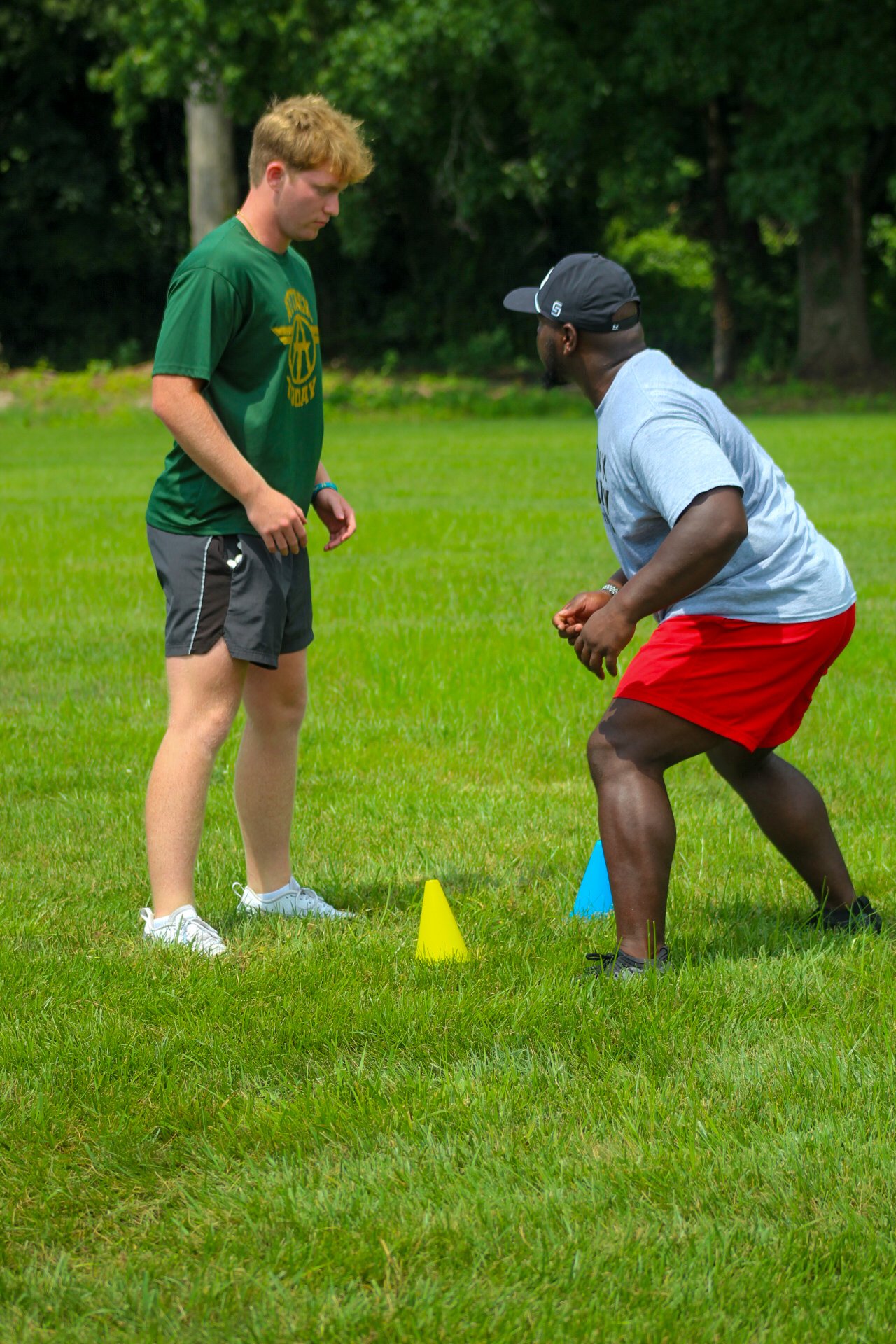 Two men are practicing football drills outdoors on a grassy field with trees in the background. One man is standing and looking down, while the other is in a crouched position between yellow and blue cones, wearing a cap and red shorts.