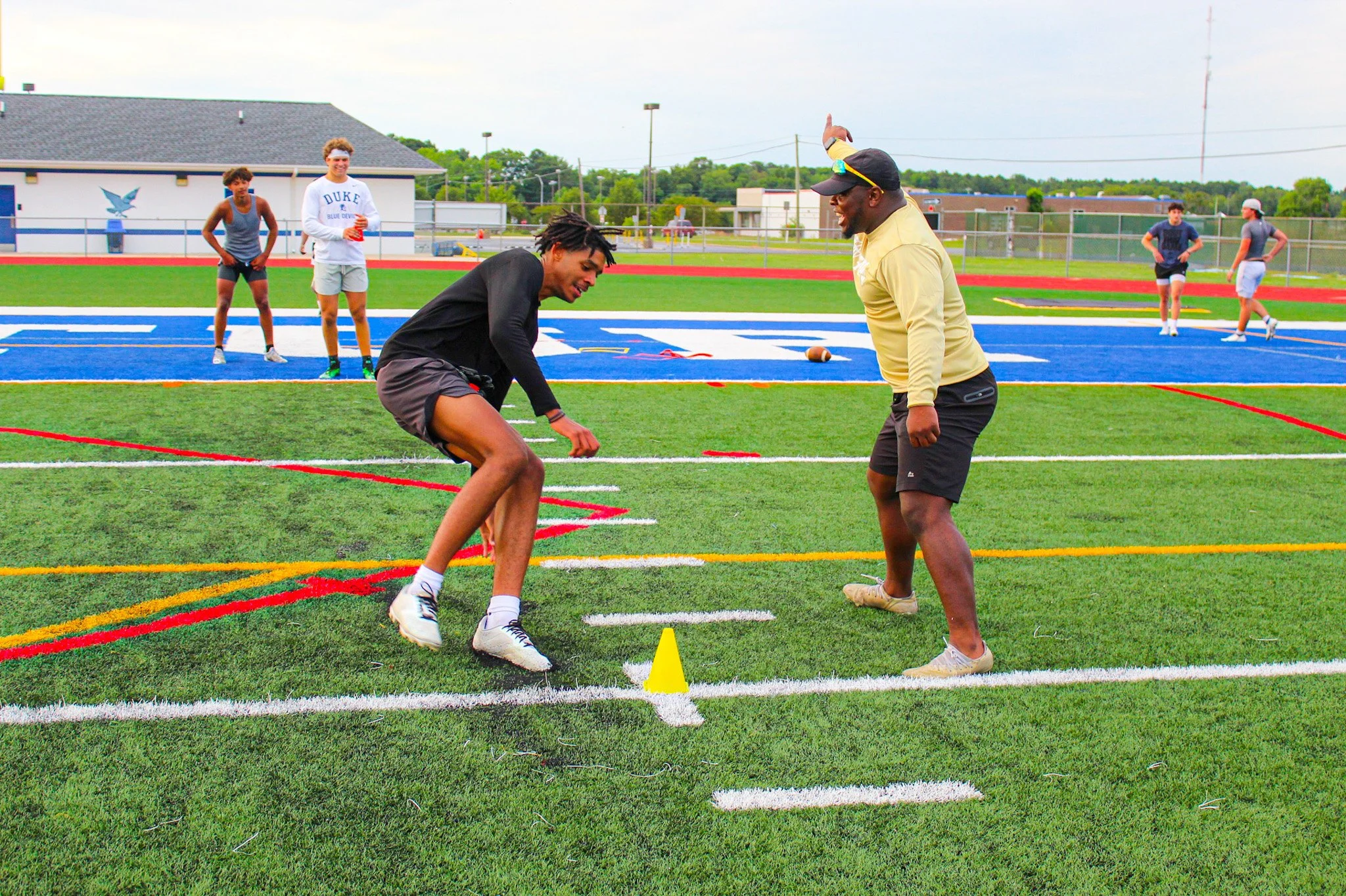 Two athletes practicing sprint drills on a track while several others observe in the background at an outdoor sports field.