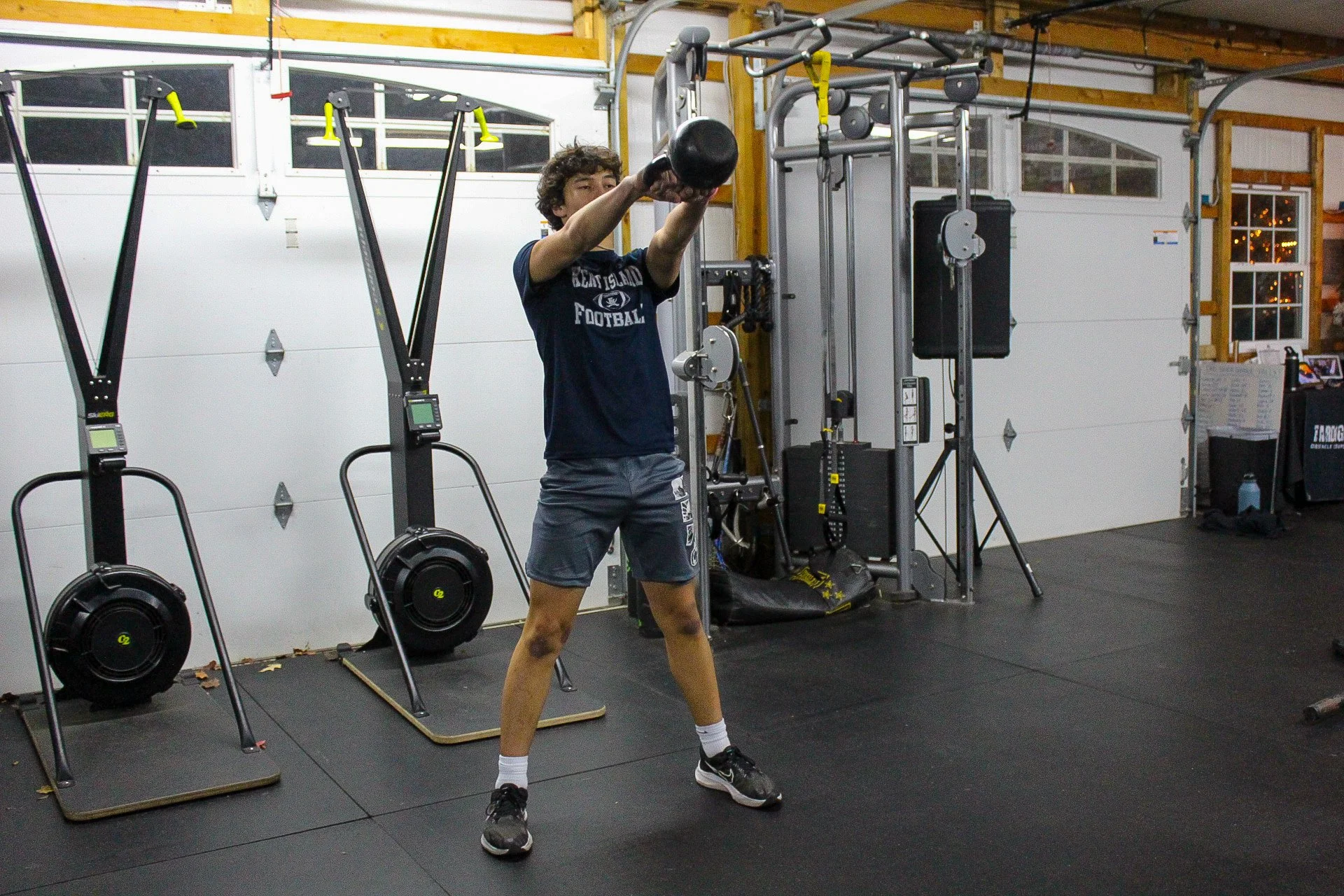 Young man performing a kettlebell swing exercise in a gym with gym equipment and a black rubber floor.