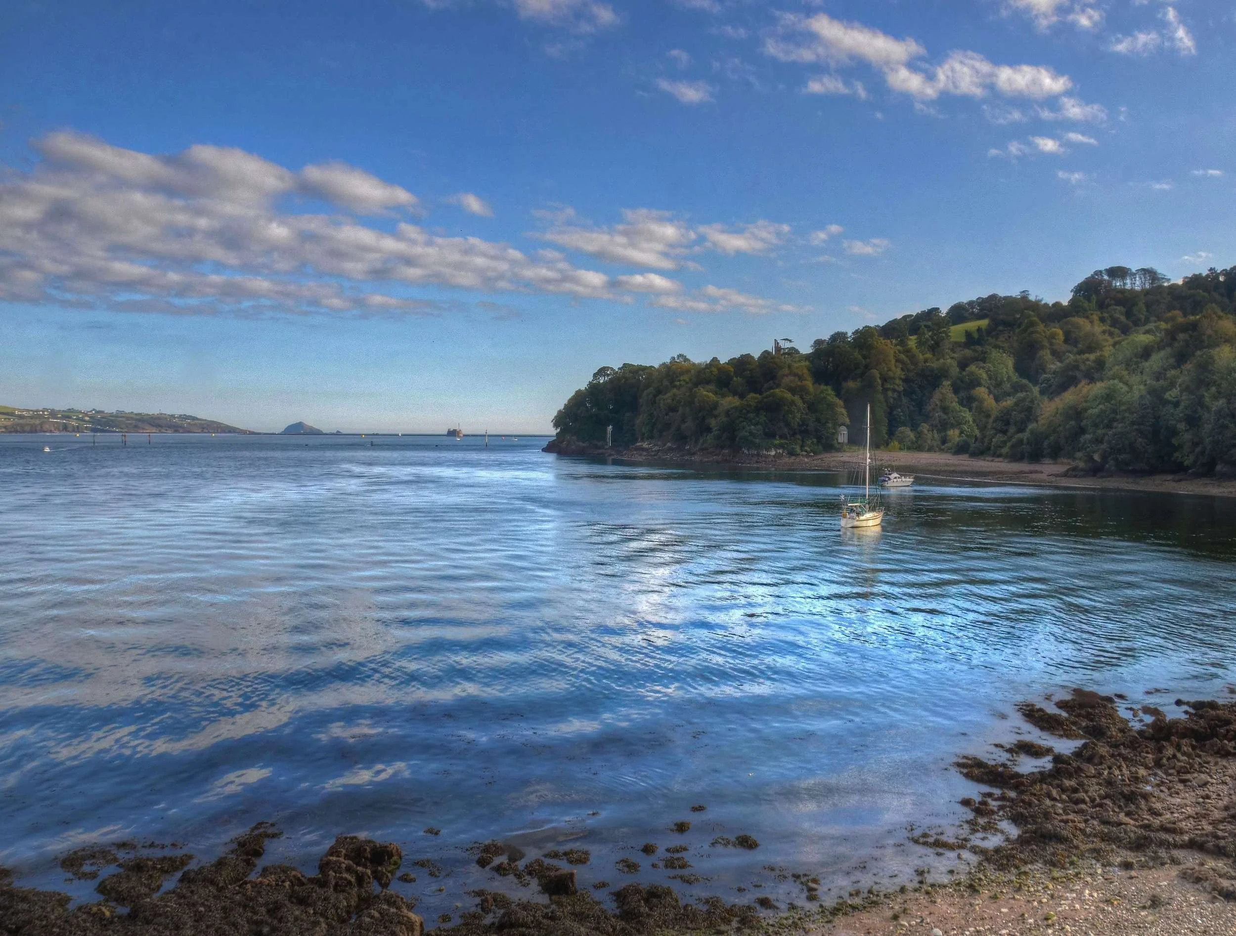Colour photo of the small beach and Milton’s Temple viewed from across still water in mellow evening light. Blue sky and fluffy clouds.