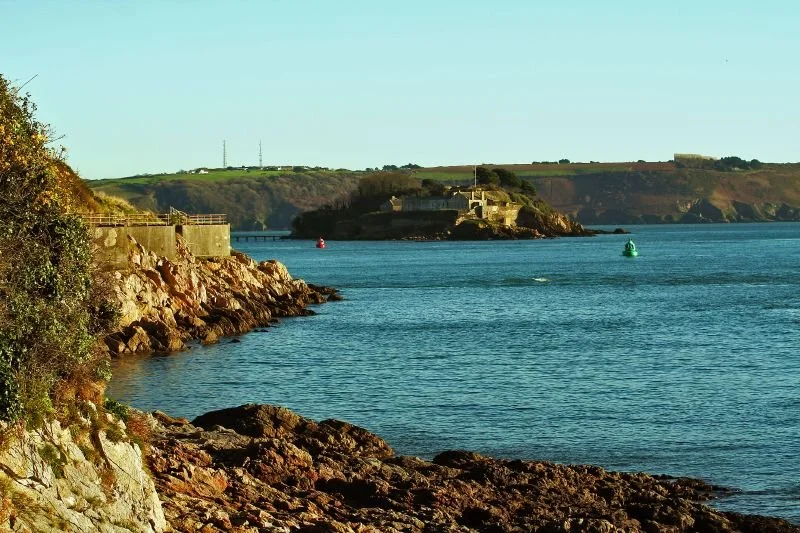 View of Drake's Island and Plymouth Sound from the country park. The sea and sky are blue. Rocky foreshore visible at front of image.